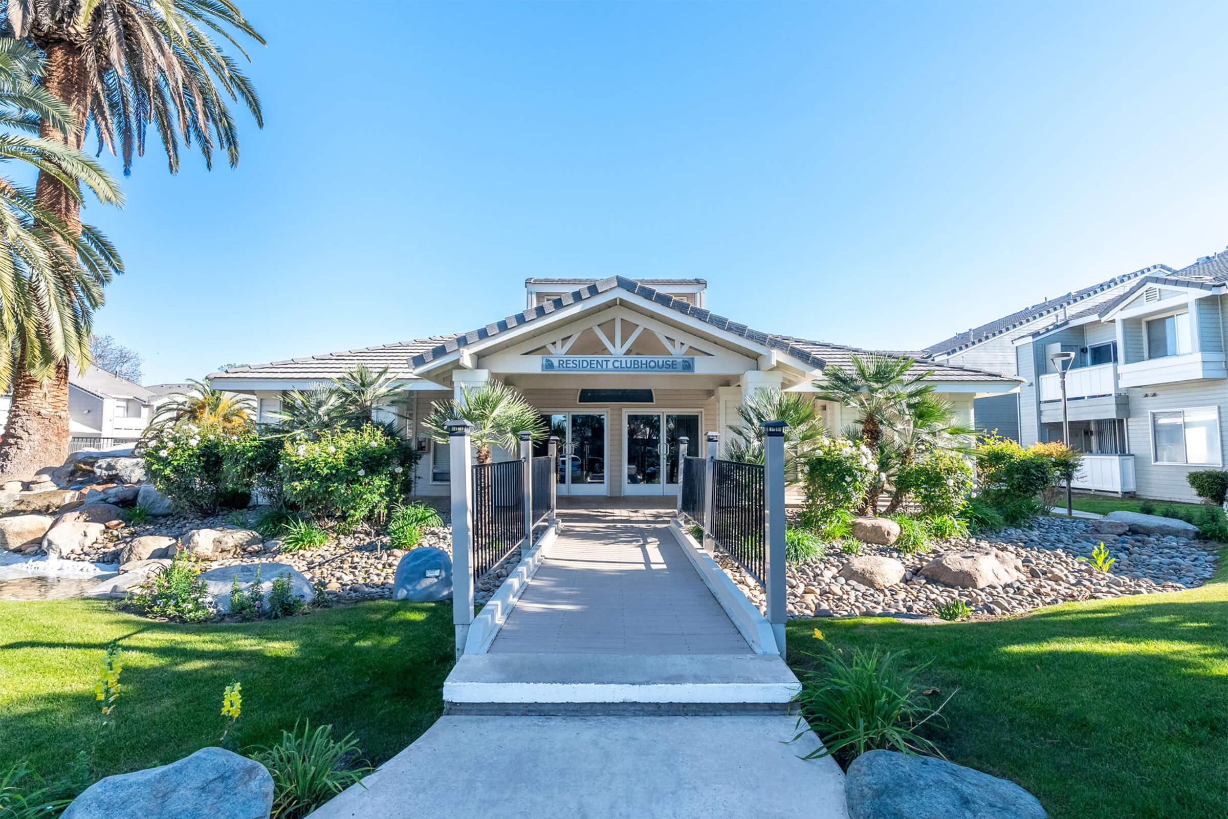 A charming residential clubhouse surrounded by palm trees and landscaped gardens. The building features a welcoming entrance with a paved pathway leading to glass doors, flanked by decorative rocks and greenery, creating a pleasant atmosphere for residents.