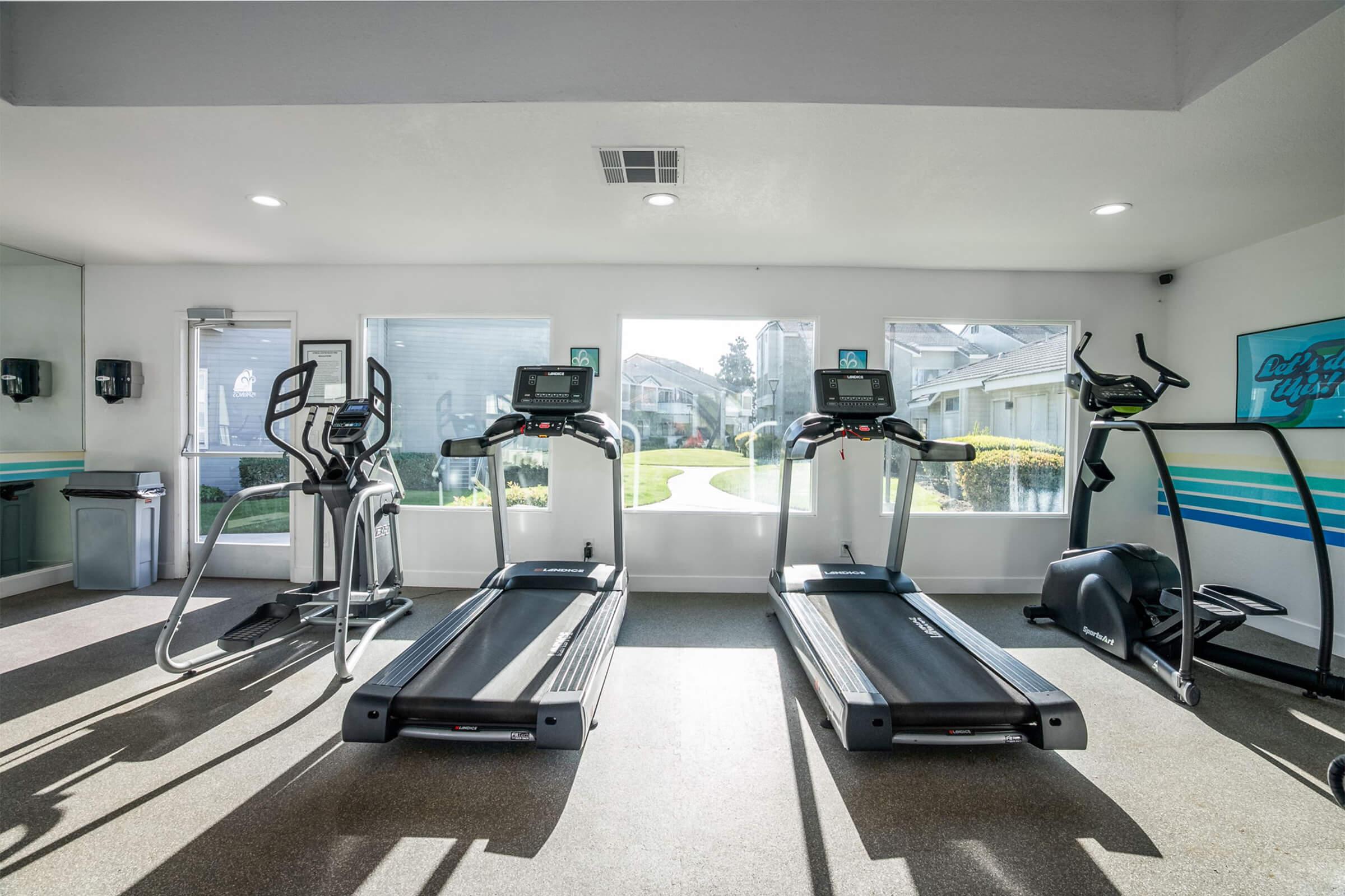 A bright and modern gym featuring three treadmills and an elliptical machine, with large windows providing natural light and a view of the outdoors. The space is clean and well-equipped, with fitness monitors on the machines and a trash bin visible in the background.