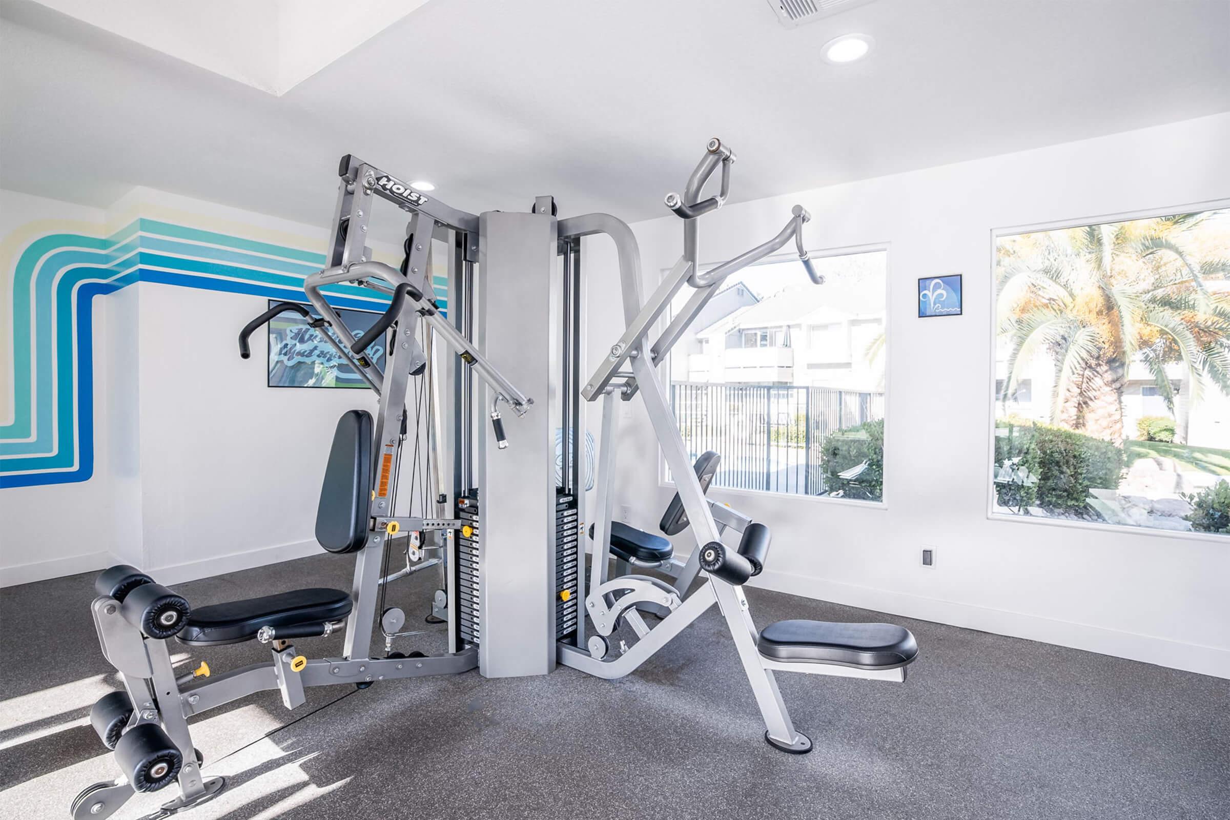 A bright and modern gym interior featuring a multi-station weight machine. The room has large windows allowing natural light, with palm trees visible outside. The walls are decorated with colorful lines, creating an inviting atmosphere for exercise and fitness activities.