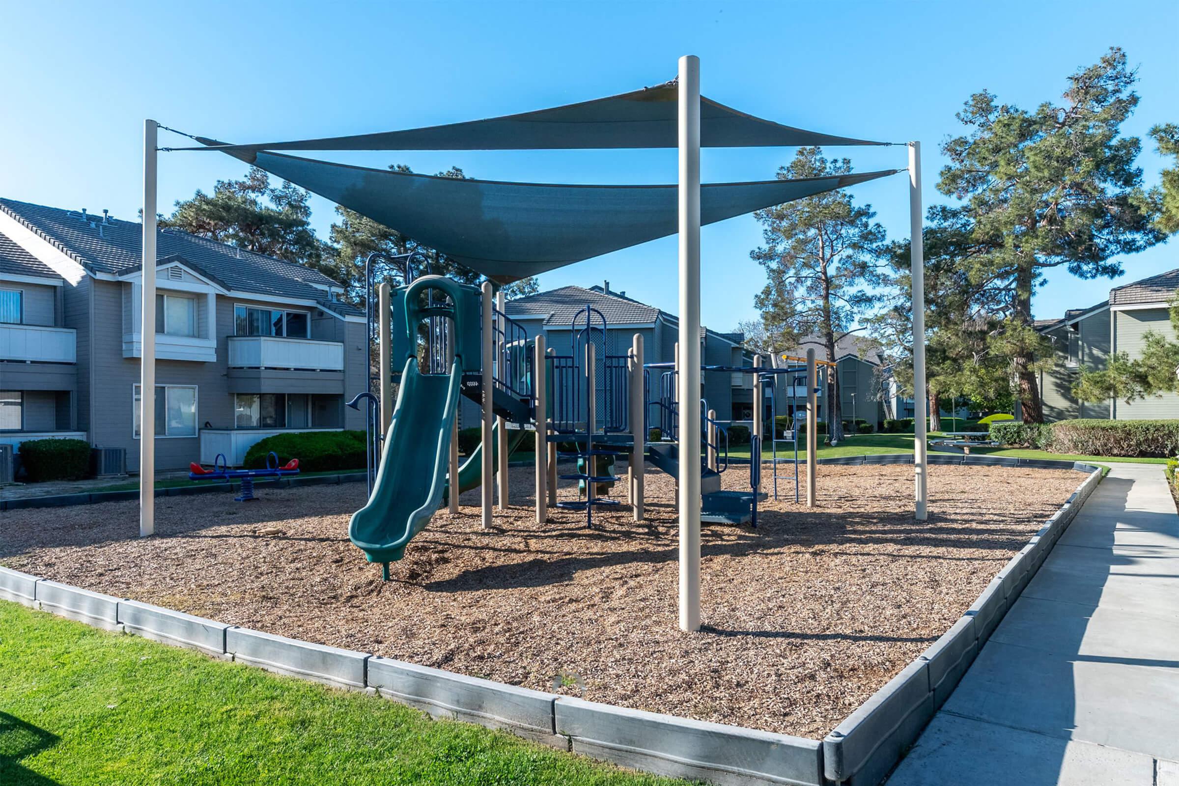 Playground area with a large green slide, climbing structures, and shaded canopies. Surrounding the playground are grassy areas and residential buildings in the background. The ground is covered with wood chips, providing a safe play surface. Clear blue sky overhead.