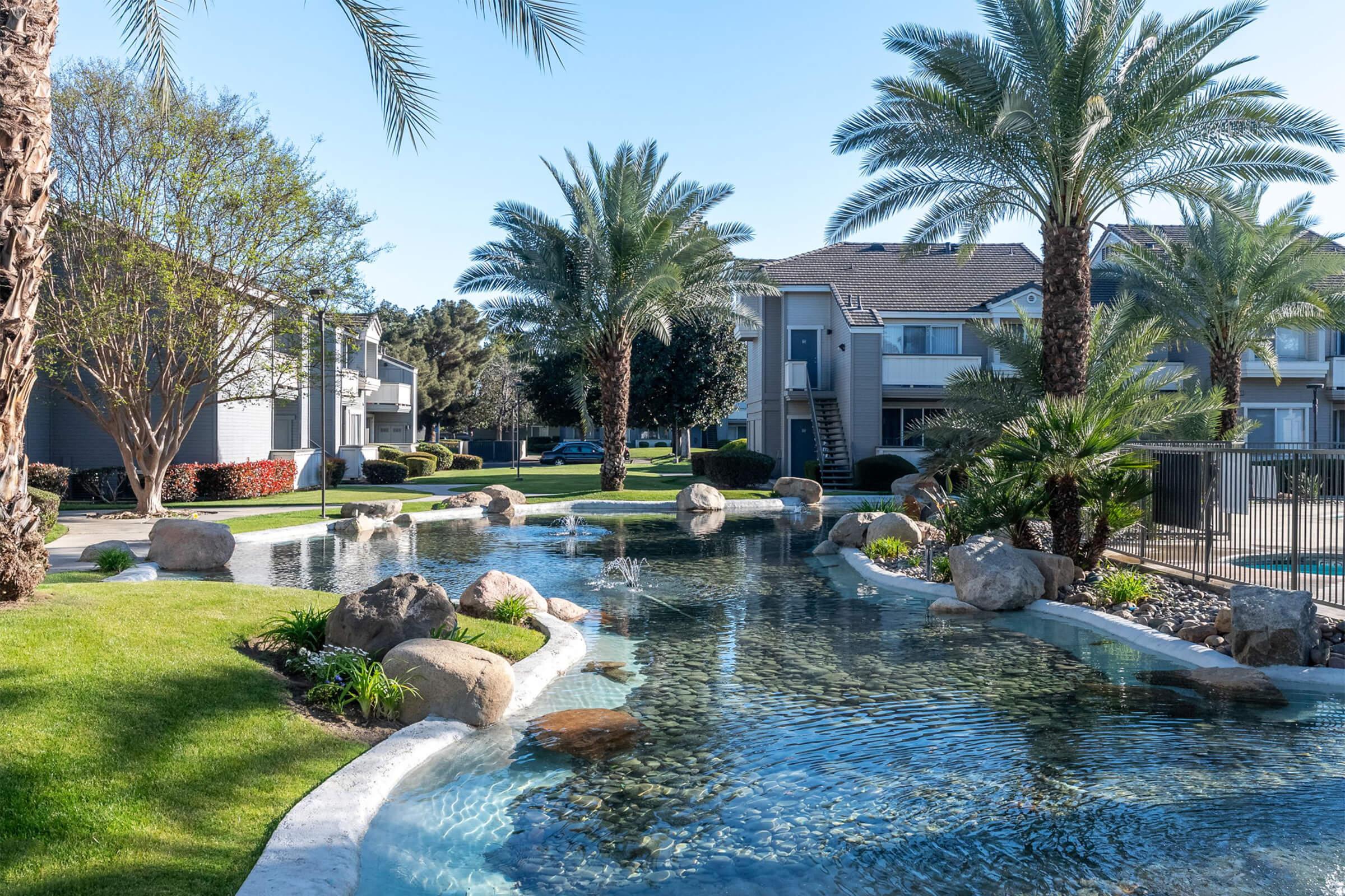 A serene landscape featuring a tranquil pond surrounded by lush greenery and palm trees. Smooth stones line the water's edge, and gentle fountains add a calming vibe. In the background, well-maintained residential buildings provide a picturesque setting for relaxation.