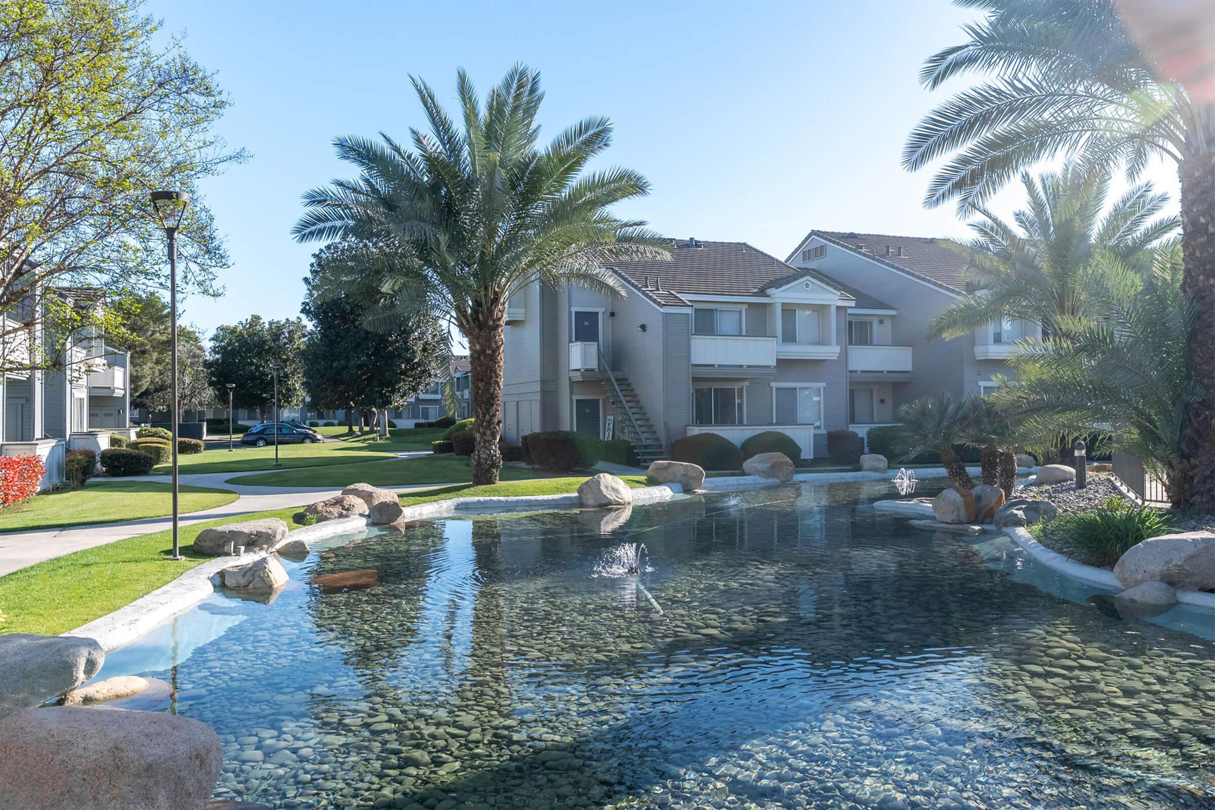 A tranquil residential area featuring a pond surrounded by rocks and palm trees. In the background, there are multi-story apartment buildings with balconies and well-maintained lawns. The scene is bathed in bright sunlight, creating a peaceful and inviting atmosphere.