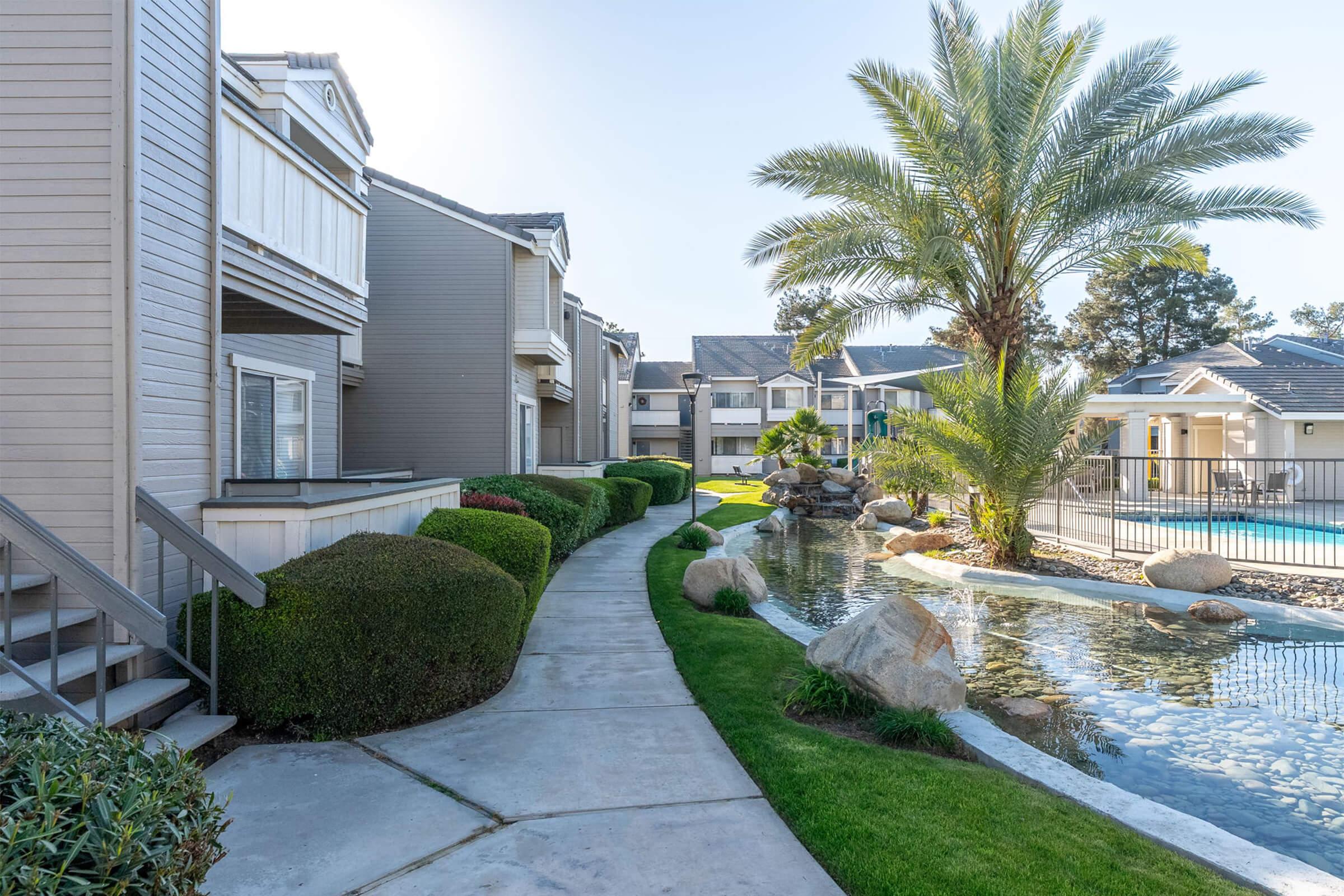 Walkway lined with greenery leading to an apartment complex, featuring a small pond with rocks and palm trees. A swimming pool is visible in the background, surrounded by well-maintained landscaping. The scene is bright and inviting, showcasing a serene residential atmosphere.