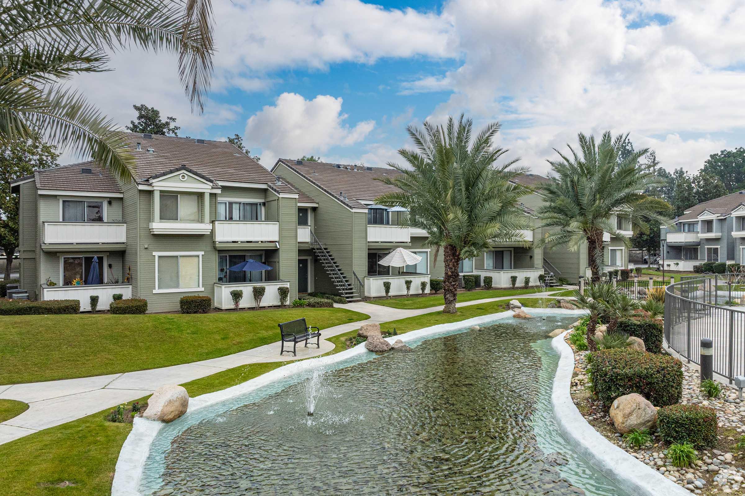 A landscaped apartment complex featuring three residential buildings, palm trees, and a winding water feature. The area is well-maintained with grassy lawns, decorative rocks, and benches for seating, under a partially cloudy sky.