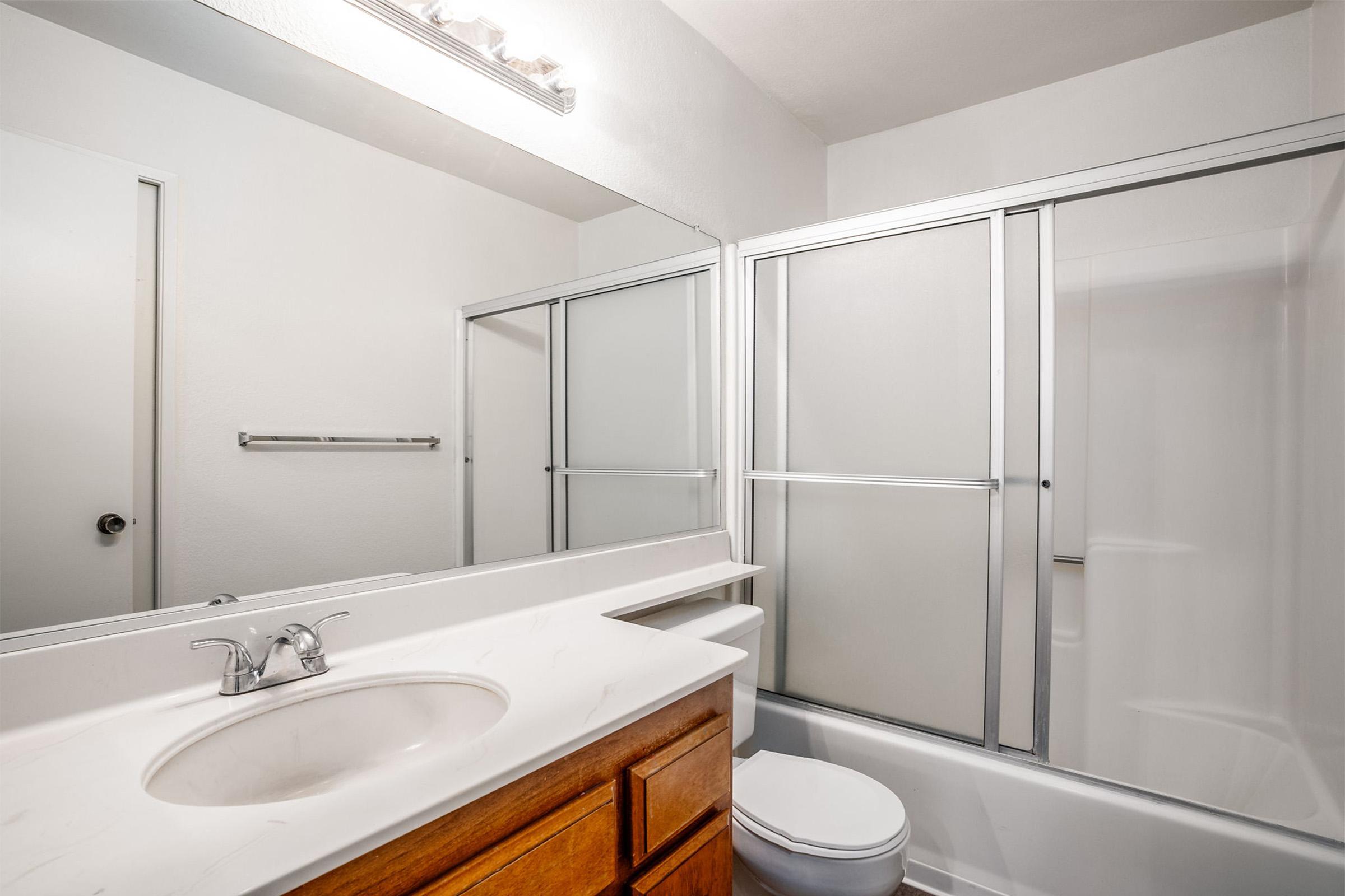 A clean, modern bathroom featuring a white countertop with a sink, a wooden cabinet below, and a large mirror above. The shower area has a glass sliding door, and a toilet is positioned beside the shower. The walls are painted white, creating a bright and airy atmosphere.
