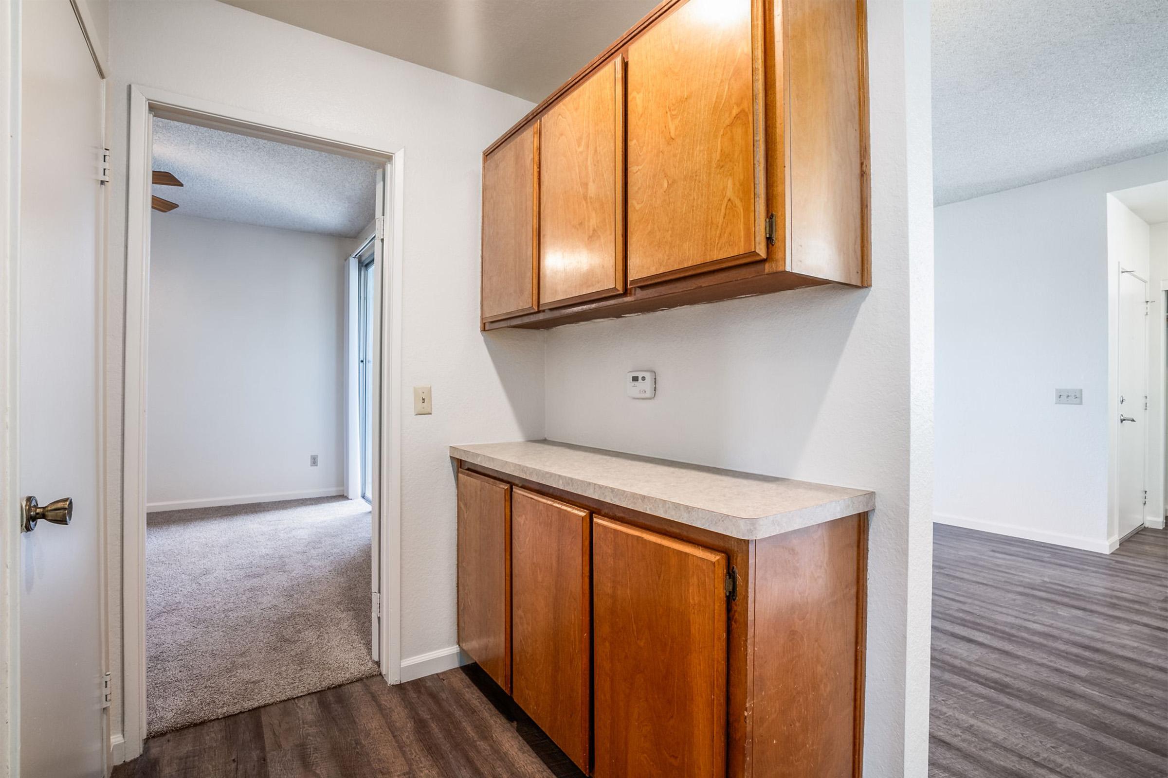 A hallway with a closed door leading to a carpeted room on the left and a kitchen area with wooden cabinets on the right. The flooring is a dark laminate, and the walls are painted white. There is a light fixture in the ceiling and a small countertop next to the cabinets.