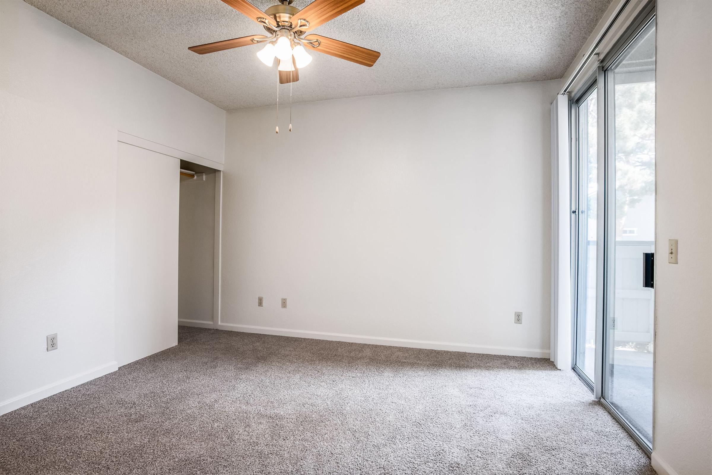 A spacious, empty room featuring light-colored walls and plush carpet. A ceiling fan with wooden blades hangs from the ceiling, while a sliding glass door opens to a balcony. A small closet is visible on the left side, adding to the room’s functionality and lightness.