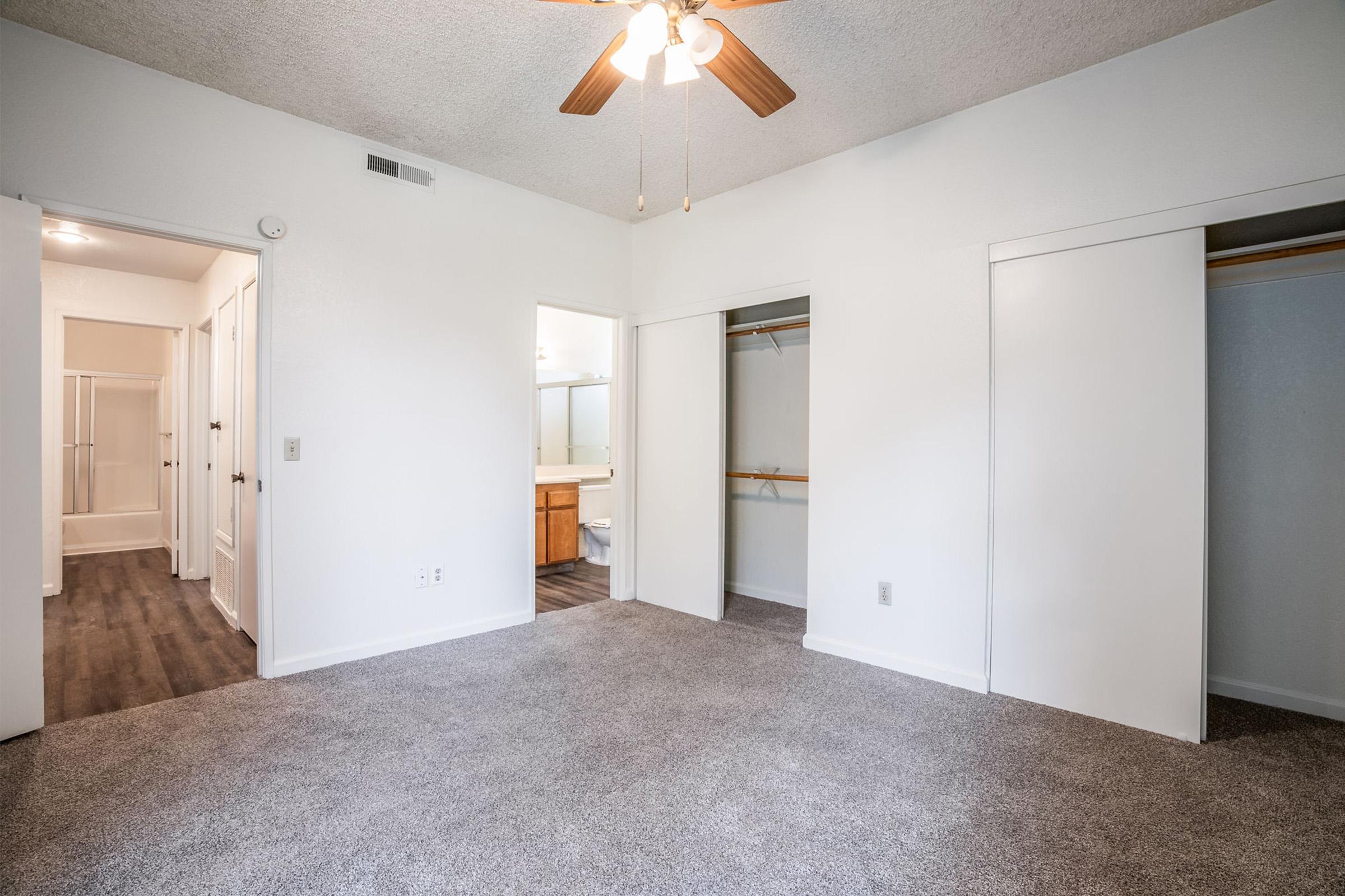 A spacious room with light-colored walls and a ceiling fan. Two doorways lead to closets and a bathroom. The floor is carpeted in a neutral shade, and there is a sense of cleanliness and openness in the space.