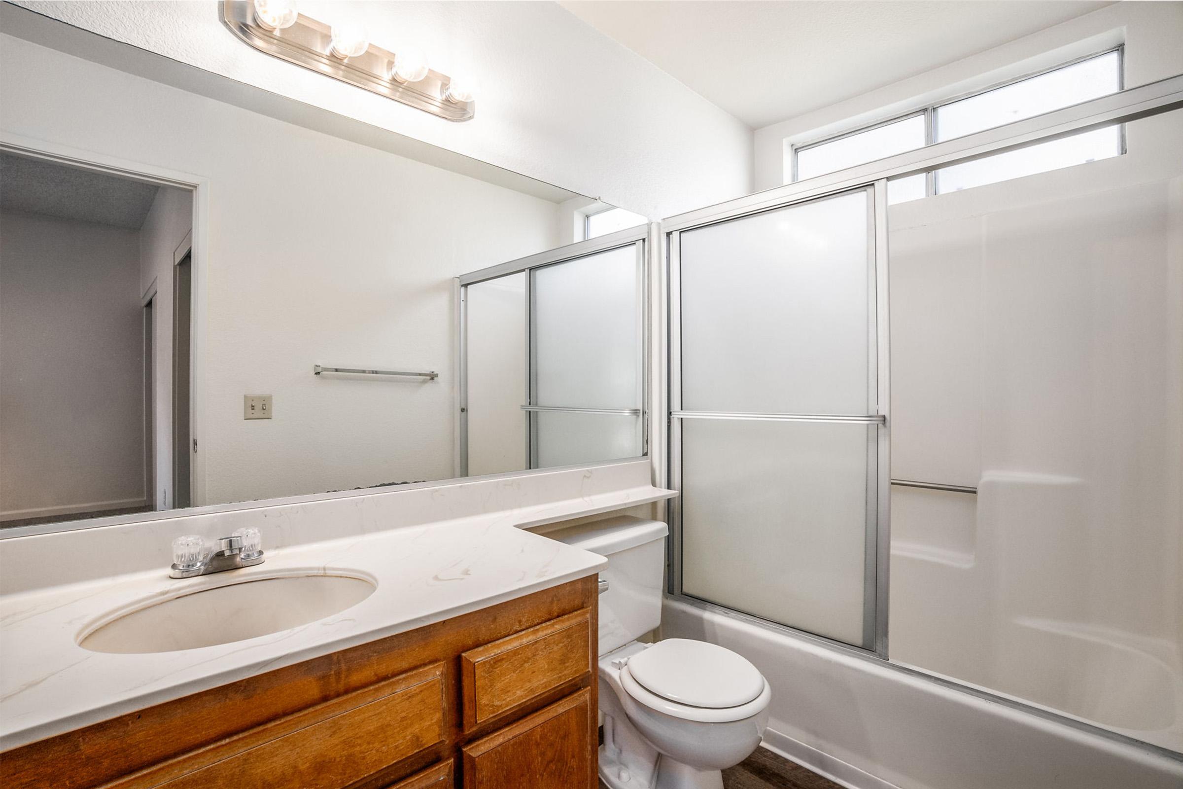 A clean and bright bathroom featuring a wooden vanity with a sink, a shower with glass doors, and a toilet. Large window providing natural light. Neutral-colored walls and flooring create a spacious feel.