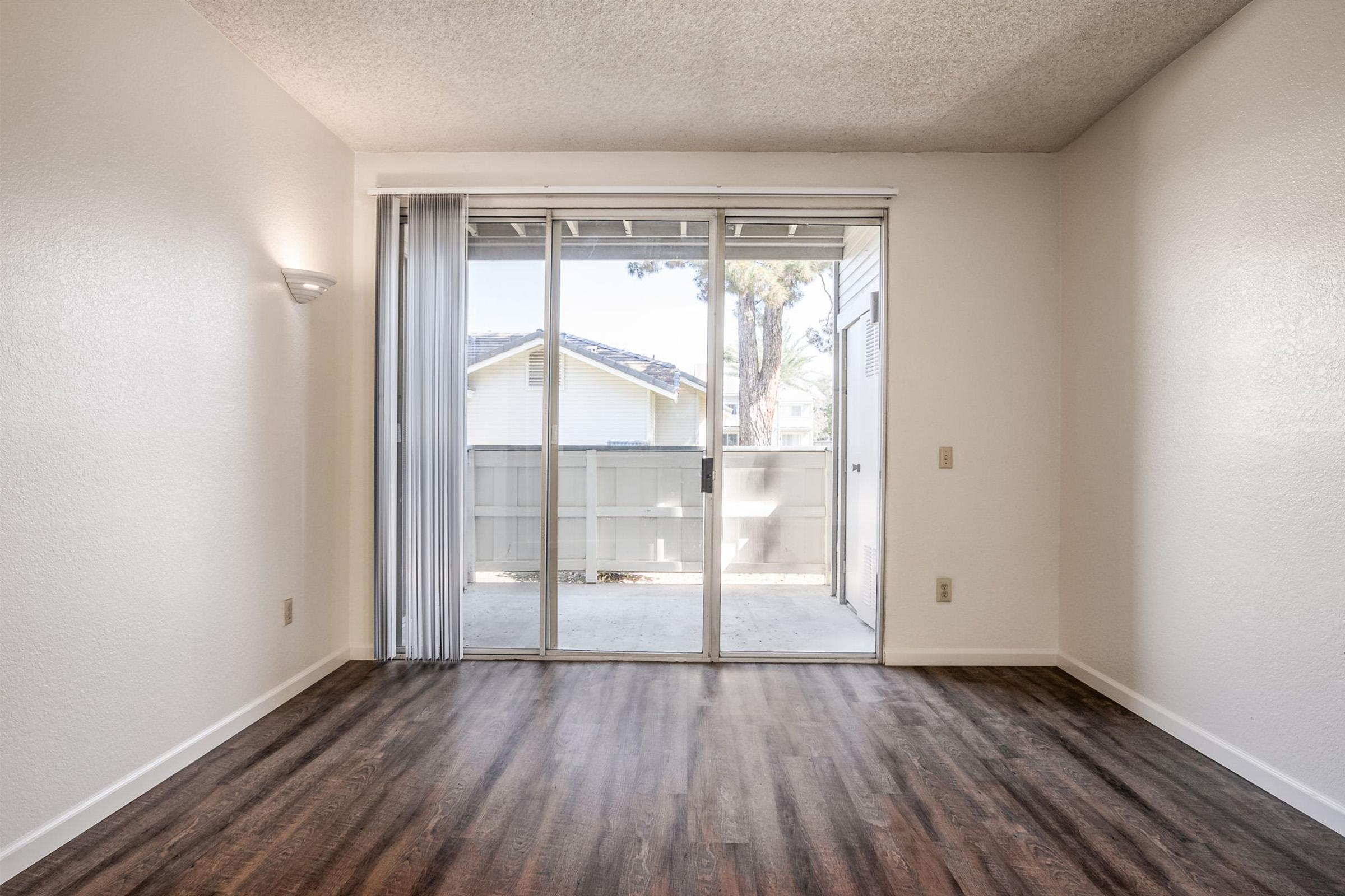 Empty room with wood-style flooring, featuring a sliding glass door leading to a patio area. Natural light filters in through the door, and there are white walls. A small wall-mounted light fixture is visible on the left side.