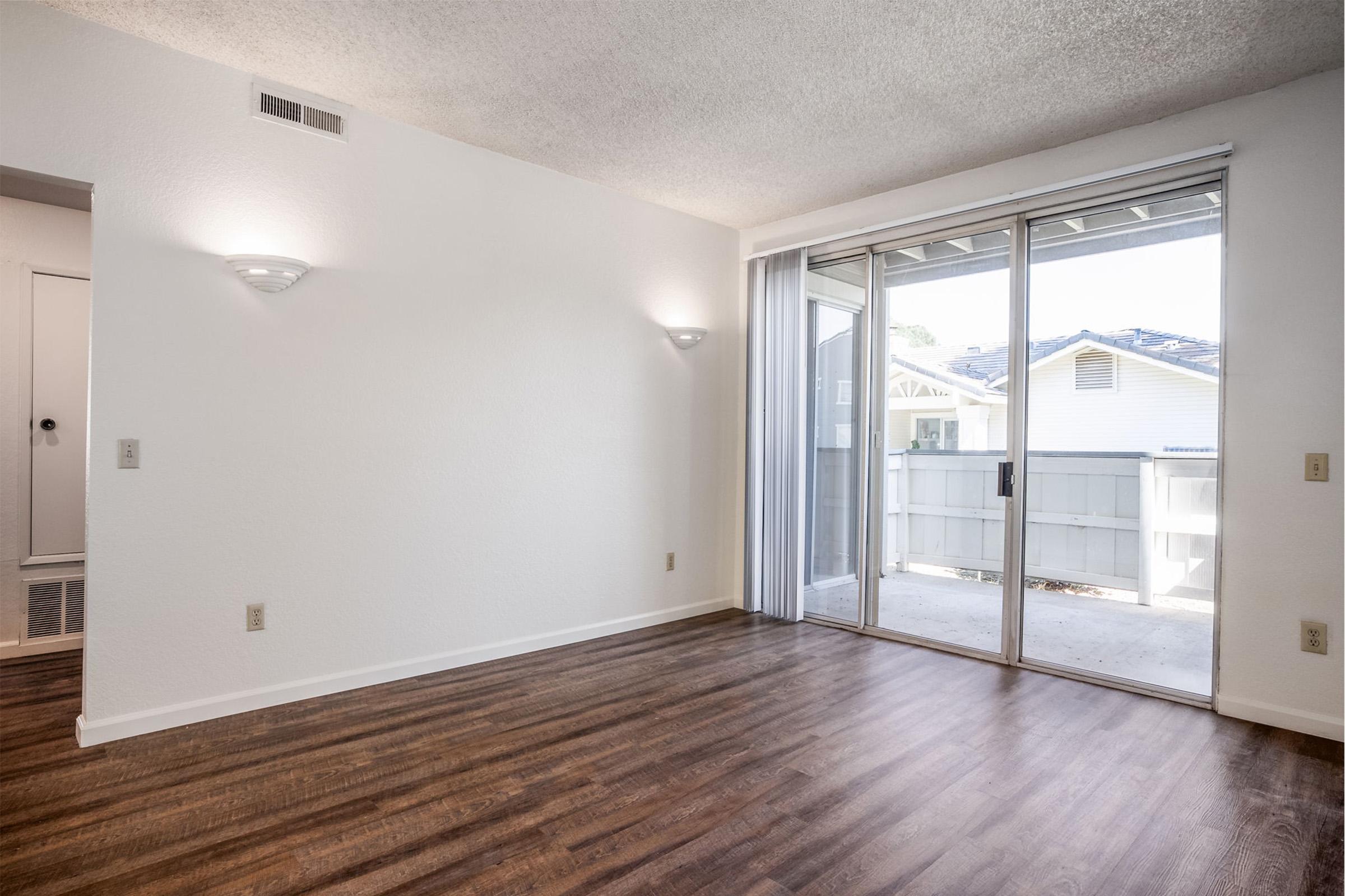 A bright, empty room with light-colored walls and wooden laminate flooring. There are two wall sconces providing gentle lighting, and a large sliding glass door leading to a balcony or patio. Natural light streams in, creating an open and inviting atmosphere.