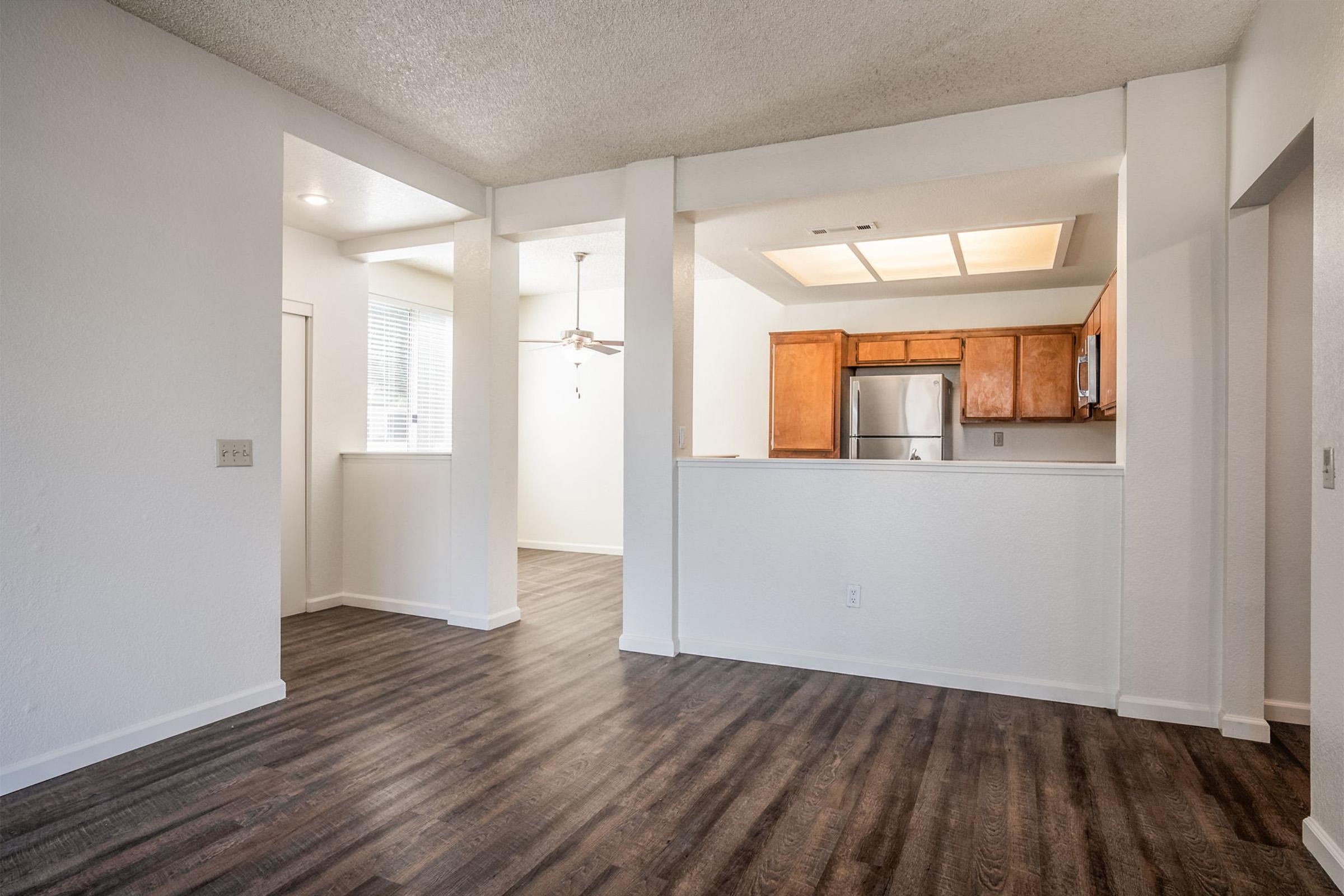 A spacious interior view of a modern apartment featuring light-colored walls, large windows, and dark wood-style flooring. The area includes a kitchen with wooden cabinetry and stainless steel appliances, and a ceiling fan is visible in the adjacent space. Natural light fills the room.