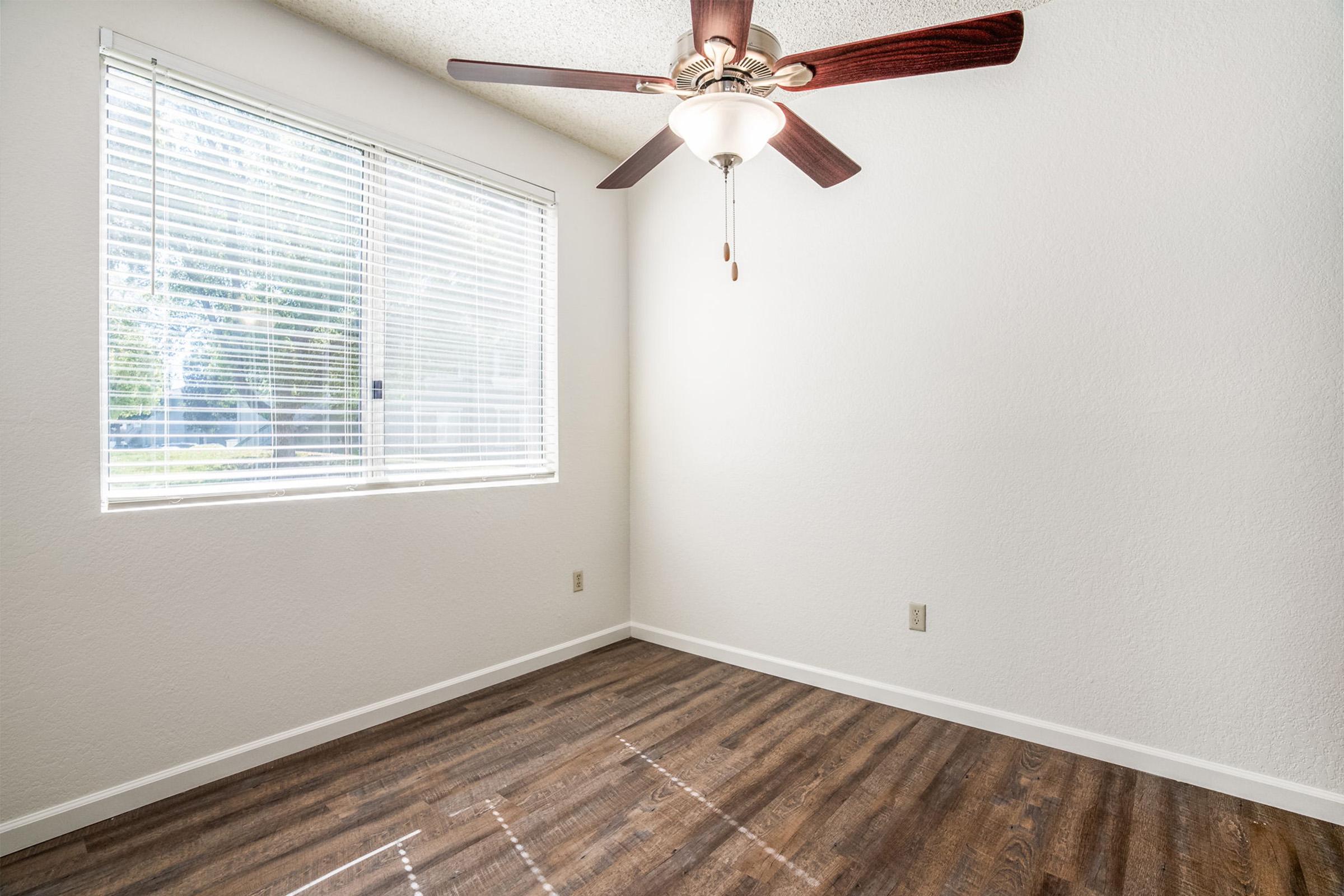 A bright and airy room with a ceiling fan and large window. The walls are painted white, and the floor features wooden laminate. Sunlight filters through the blinds, illuminating the spacious feel of the room, which is unfurnished and ready for personalization.