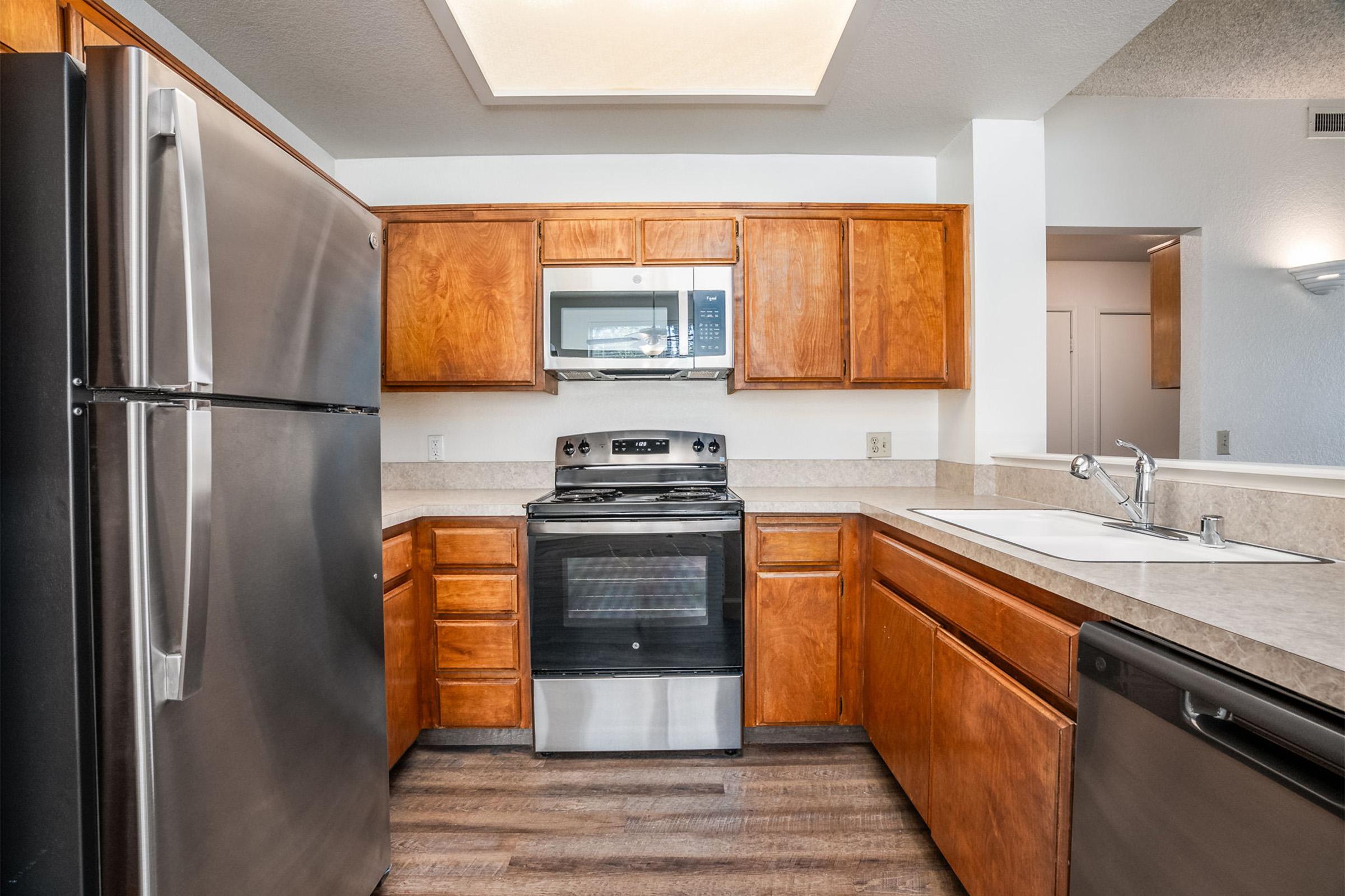 Modern kitchen featuring stainless steel appliances, including a refrigerator, oven, and microwave. The cabinets are a warm wood finish, and the countertops are light-colored with a textured surface. The space has a spacious layout with a sink and dishwasher, complemented by a soft overhead light.