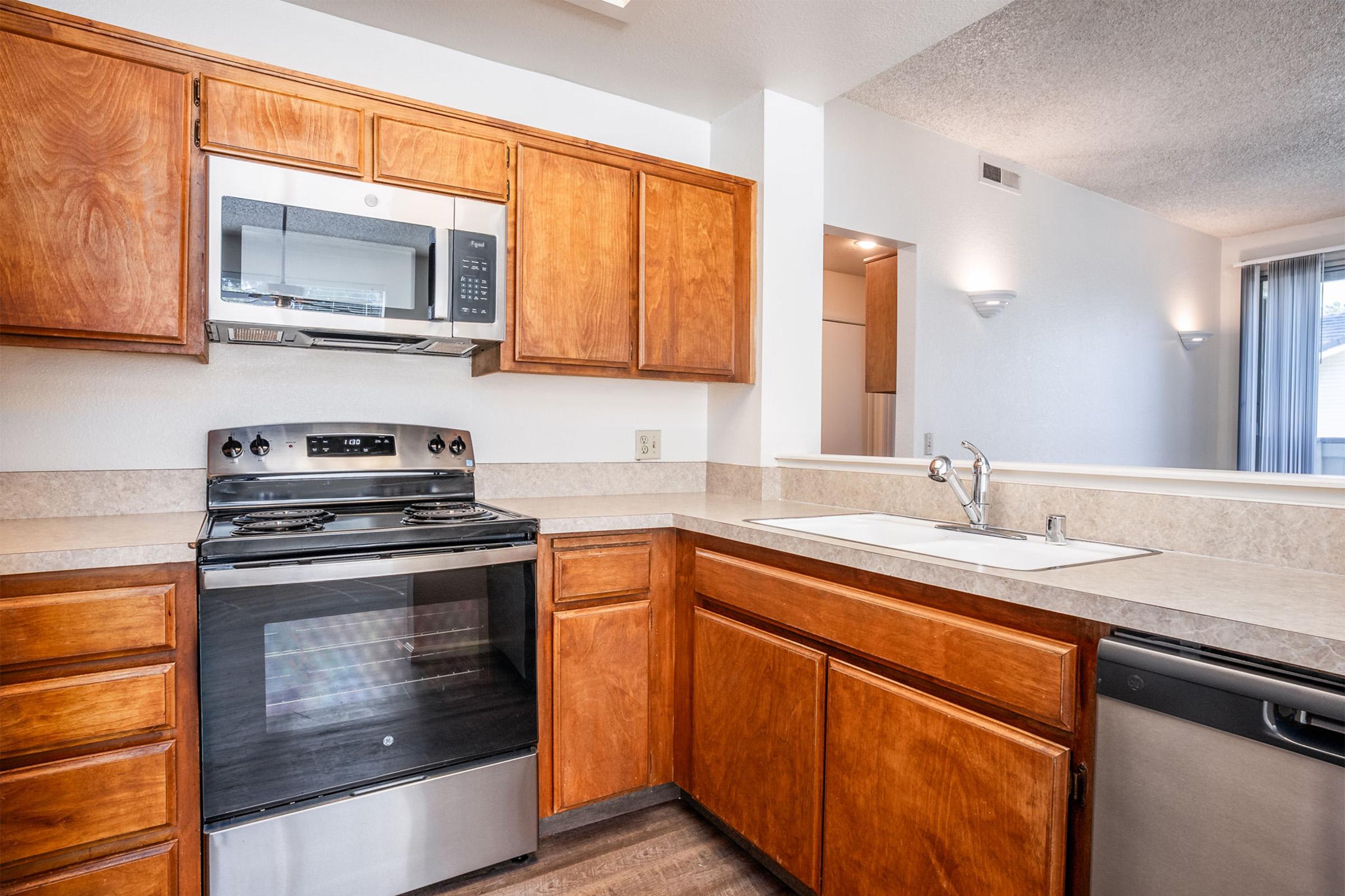 Modern kitchen featuring wooden cabinets, stainless steel appliances including a microwave and oven, a sink with a faucet, and granite countertops. Natural light fills the space, creating a warm atmosphere. The layout is open, connecting to a bright living area with sliding glass doors.