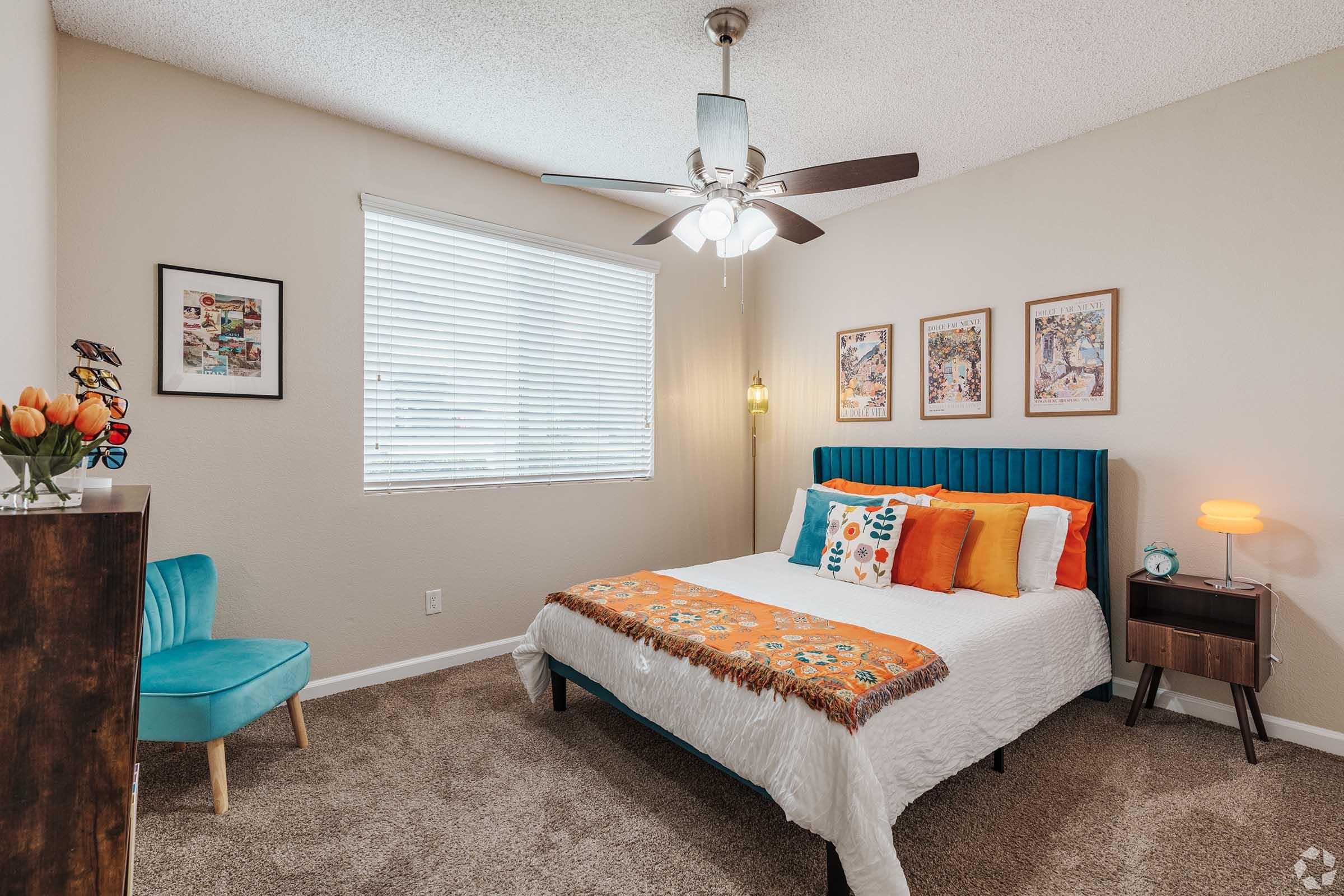 A cozy bedroom featuring a blue accent wall with a bed dressed in white and colorful throw pillows, a wooden bedside table with a lamp, and a teal chair in the corner. The walls are adorned with framed art, and a ceiling fan with light fixtures completes the inviting atmosphere.