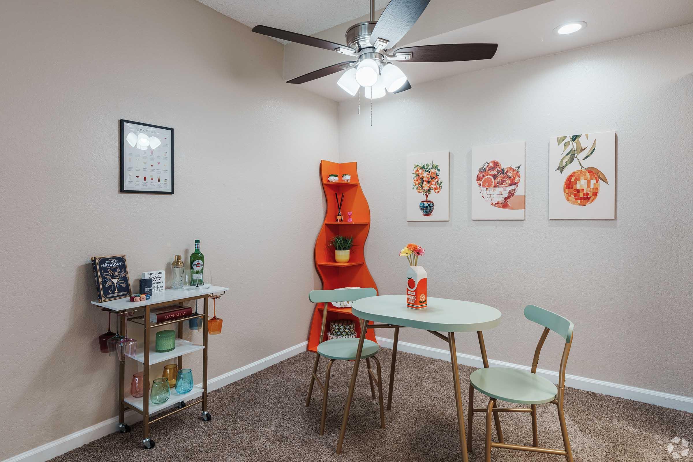 A cozy dining area featuring a round mint green table and two light green chairs. There's a small cart with drinks, a wall-mounted shelf displaying decorative items, and three art pieces on the wall. A ceiling fan with lights and neutral-colored walls create a warm atmosphere.