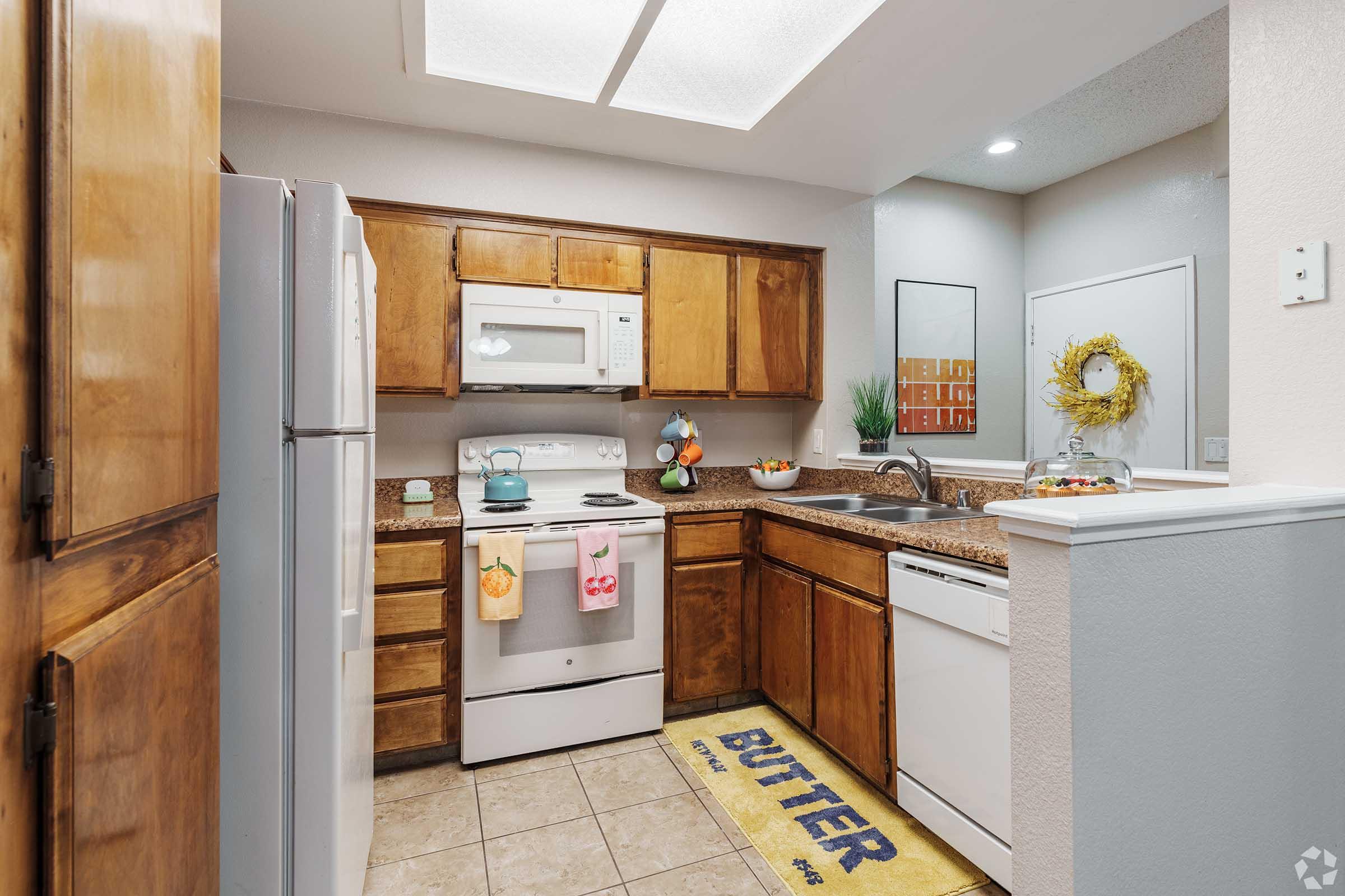 A modern kitchen featuring wooden cabinets, a granite countertop, and stainless steel appliances. The kitchen is equipped with a microwave, oven, and dishwasher, and includes a decorative rug that reads "BUTTER." Natural light enters through a square ceiling light, enhancing the inviting atmosphere.
