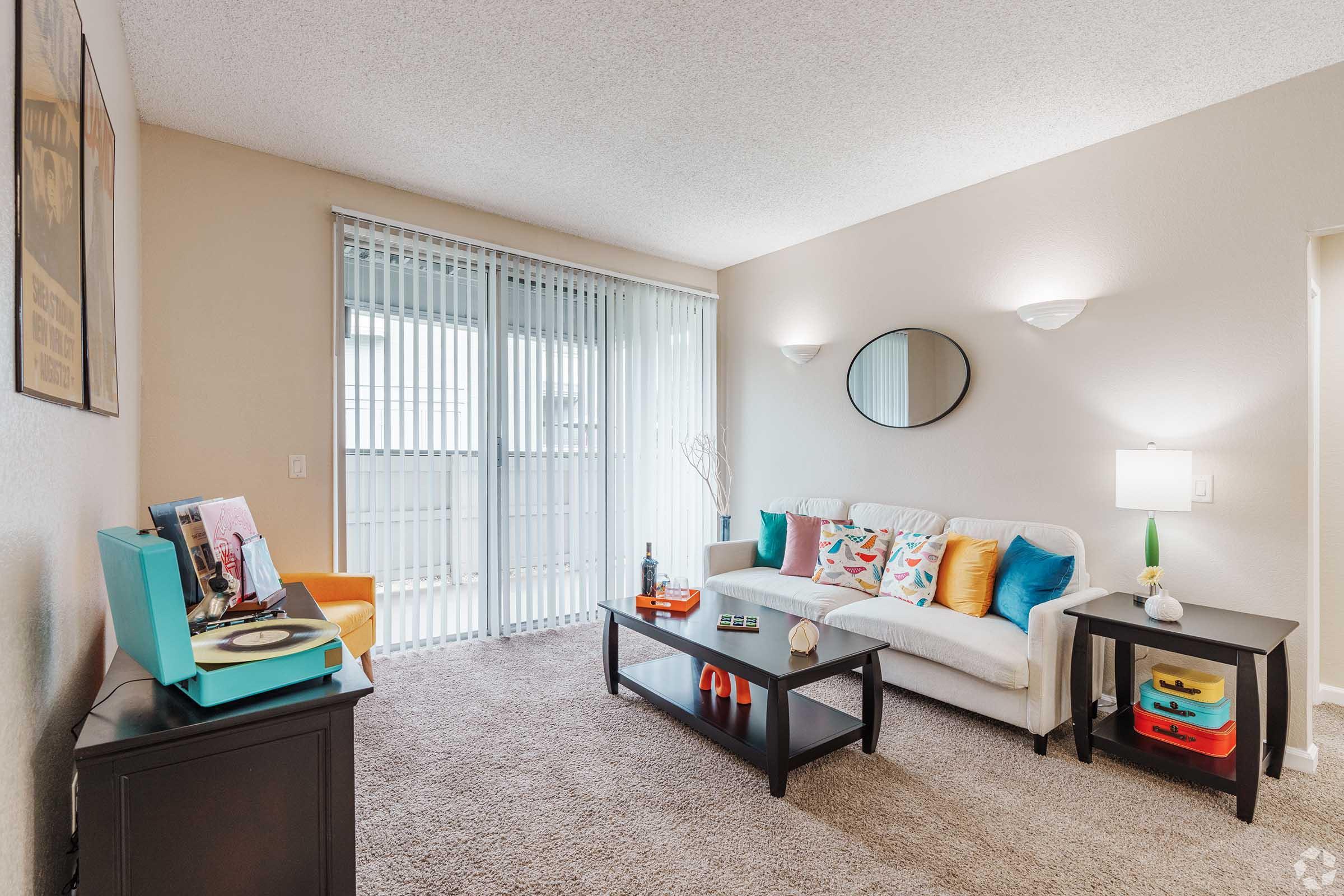 Bright and cozy living room featuring a white sectional sofa adorned with colorful throw pillows. A coffee table sits in front, with a record player on it. Large sliding glass doors allow natural light, and there are decorative wall art pieces. The carpeted floor adds warmth to the space.