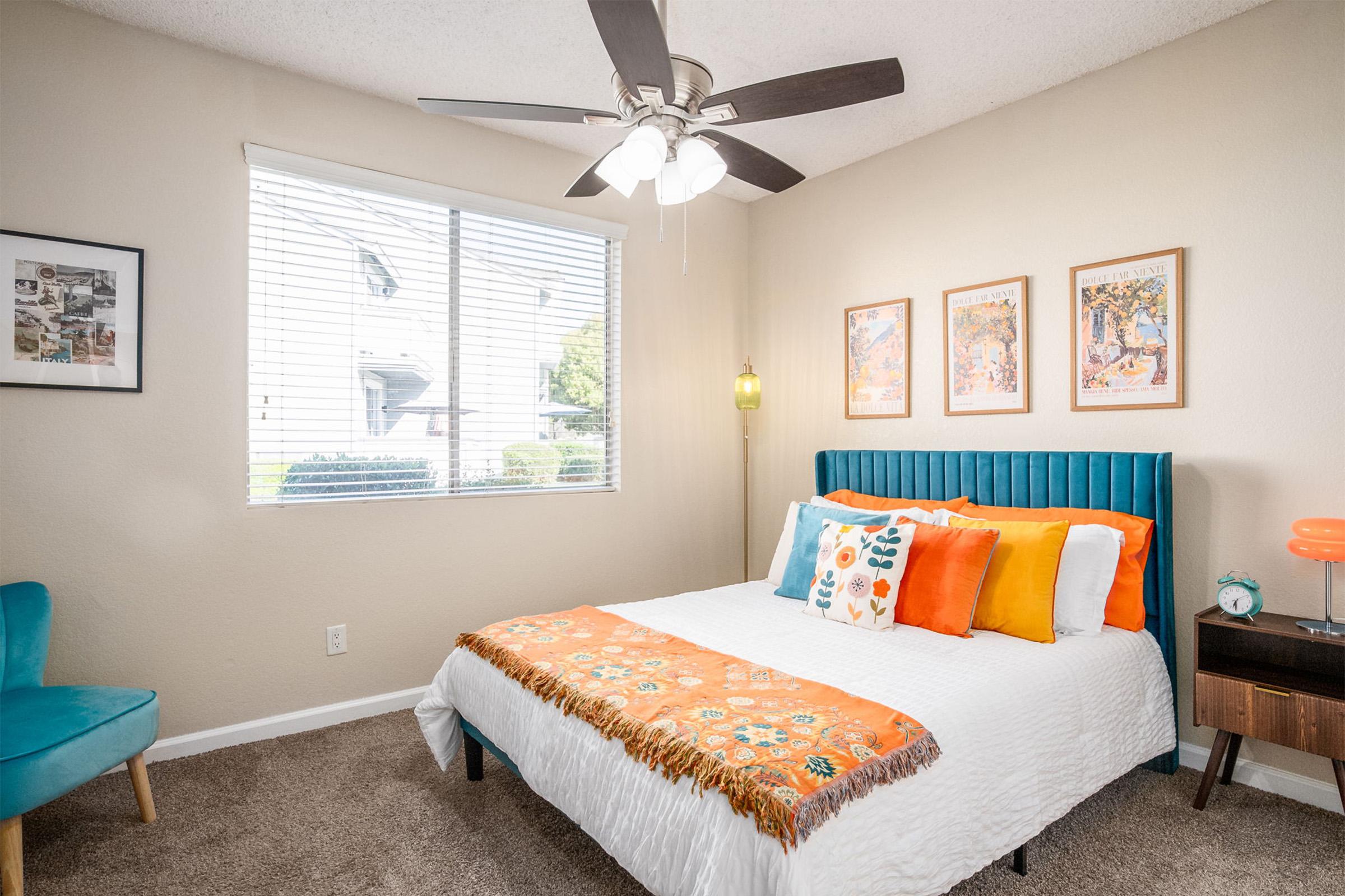 A cozy bedroom featuring a double bed with colorful pillows and a decorative throw blanket. There's a blue accent chair, a bedside table with a lamp, and framed artwork on the walls. Large windows allow natural light to fill the space, showcasing a pleasant view outside.