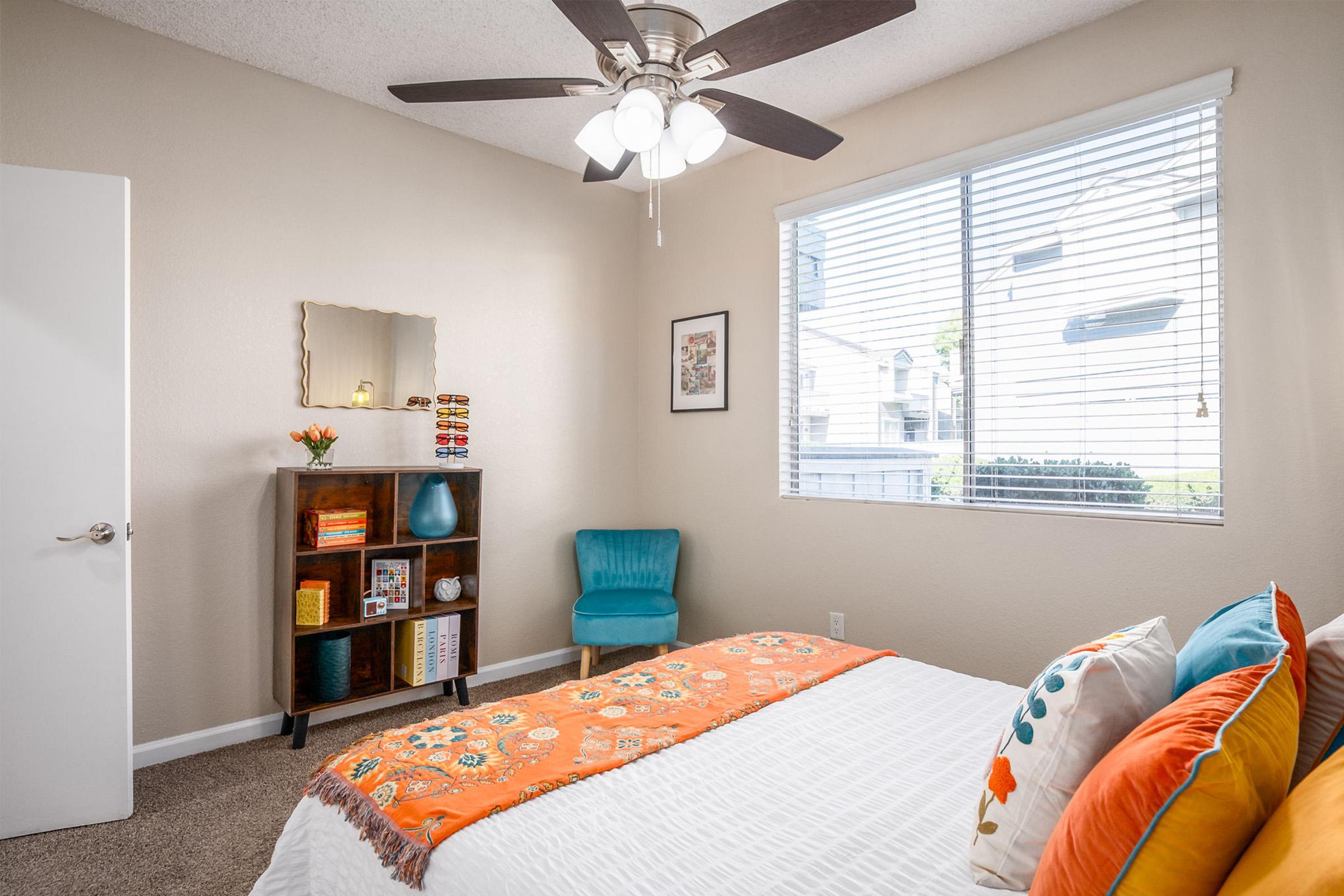 Brightly lit bedroom featuring a white bed with colorful pillows and an orange throw blanket. A small bookshelf holds decorative items and books. A blue accent chair is positioned beside the bed, and a wall-mounted mirror reflects natural light coming through a large window with blinds.