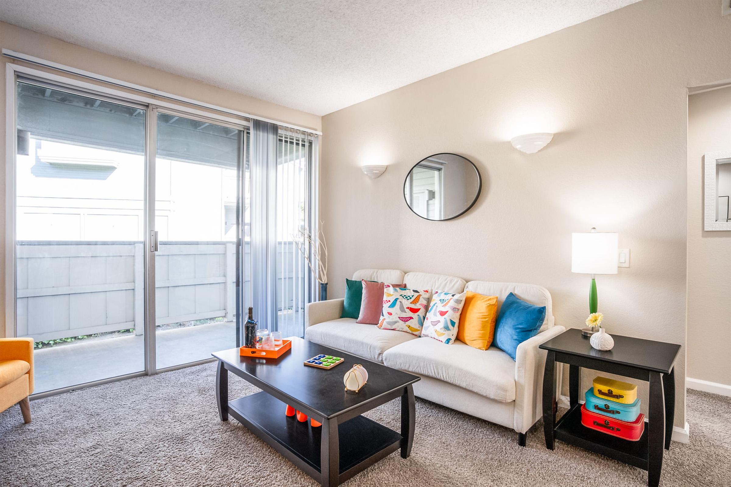 Bright and inviting living room featuring a white sofa with colorful cushions, a black coffee table with decorative items, and a lamp. Large sliding glass doors provide natural light and a view outside. A cozy orange armchair complements the space, enhancing the warm, welcoming atmosphere.