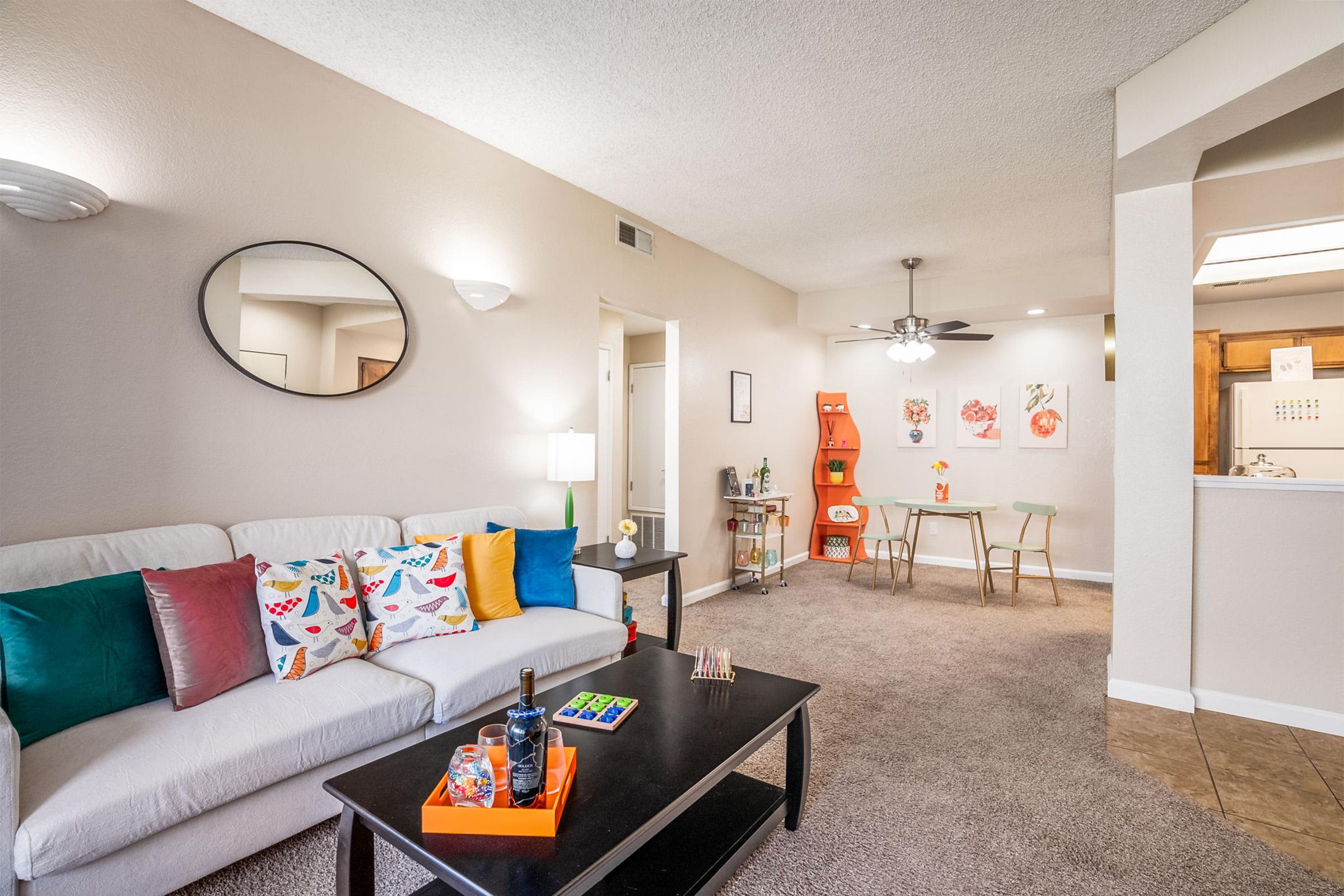 Cozy living room featuring a white sofa adorned with colorful pillows, a black coffee table, and a lamp. A small dining area with a round table and chairs is visible, along with decorative wall art. The space has a light, inviting atmosphere with neutral walls and a ceiling fan.