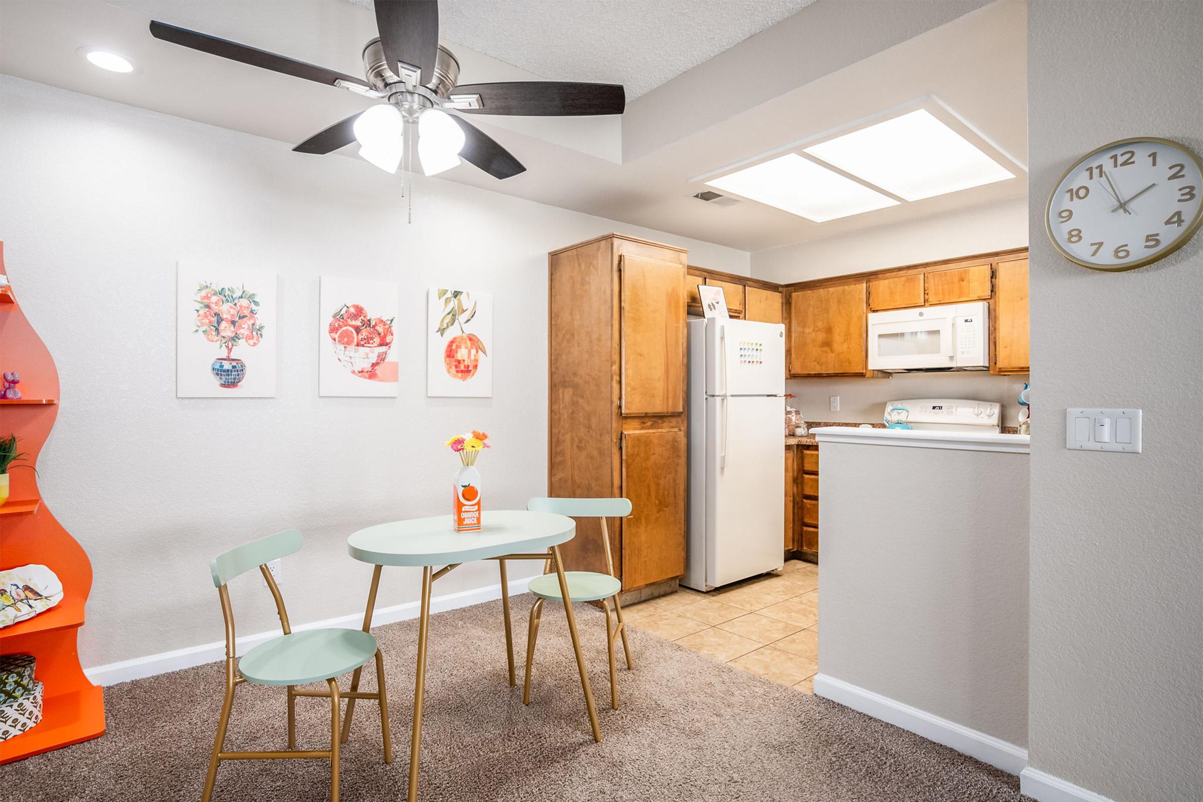 A cozy dining area featuring a round light green table with two matching chairs, a vase of flowers as table decor, and colorful artwork on the walls. In the background, a modern kitchen with wooden cabinets, white appliances, and a ceiling fan can be seen. The floor is carpeted, creating a warm atmosphere.