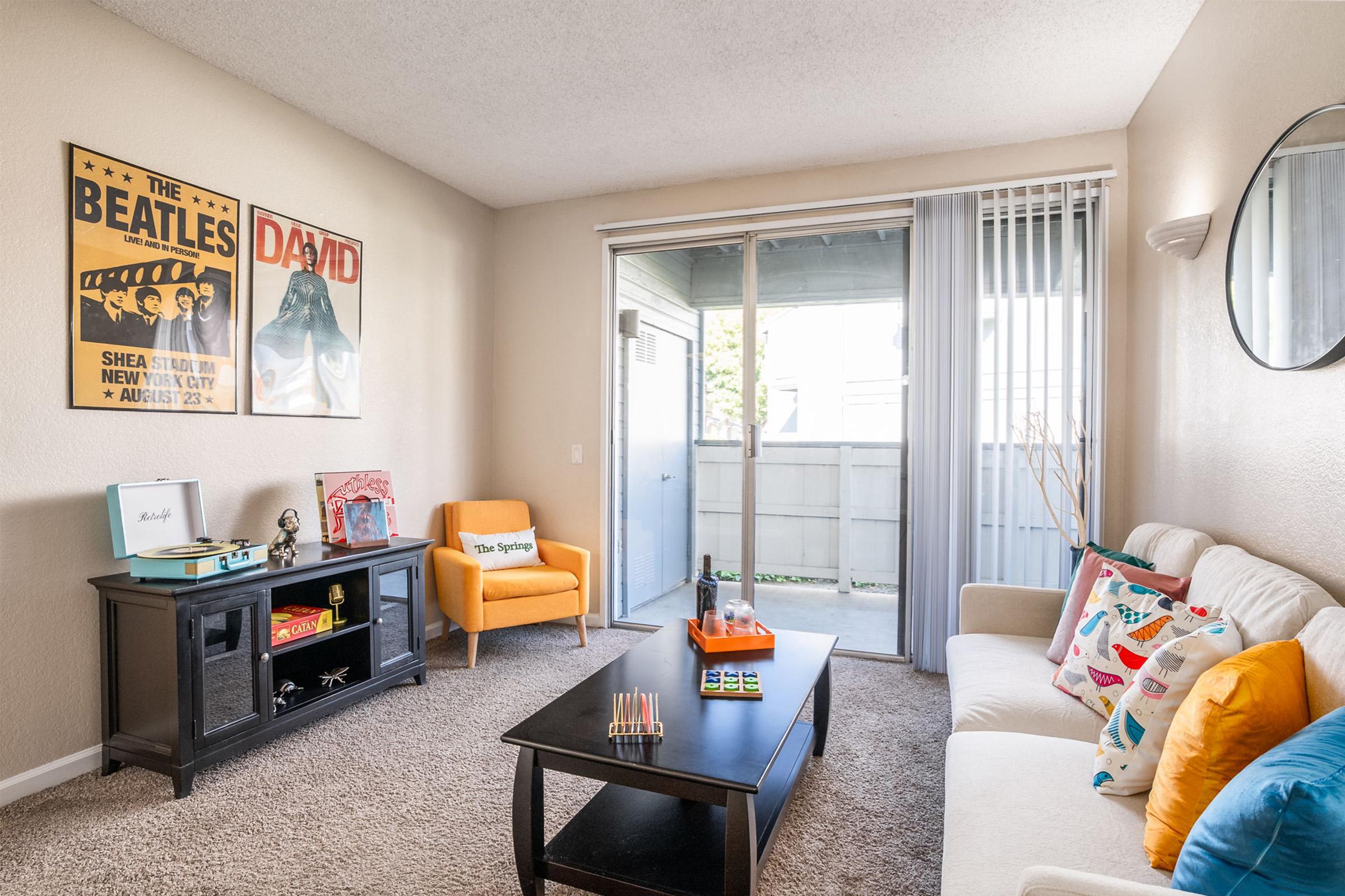 A cozy living room featuring a small white sofa with colorful cushions, a coffee table with games on it, and a black TV stand with decorative items. An orange armchair adds a pop of color, while artwork of The Beatles and David Bowie adorns the walls. The room is well-lit with large windows and sliding glass doors.