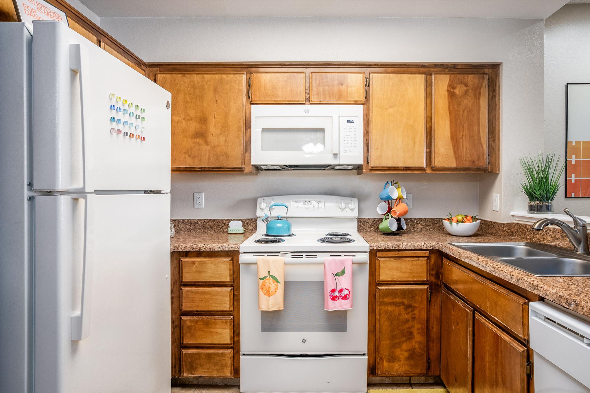 A clean and modern kitchen featuring wooden cabinetry, a white refrigerator with colorful magnets, a white stove with a microwave above, and a stainless steel sink. Decorative items include a teal kettle, colorful dish towels, and a fruit bowl on the counter. Natural light enhances the inviting atmosphere.