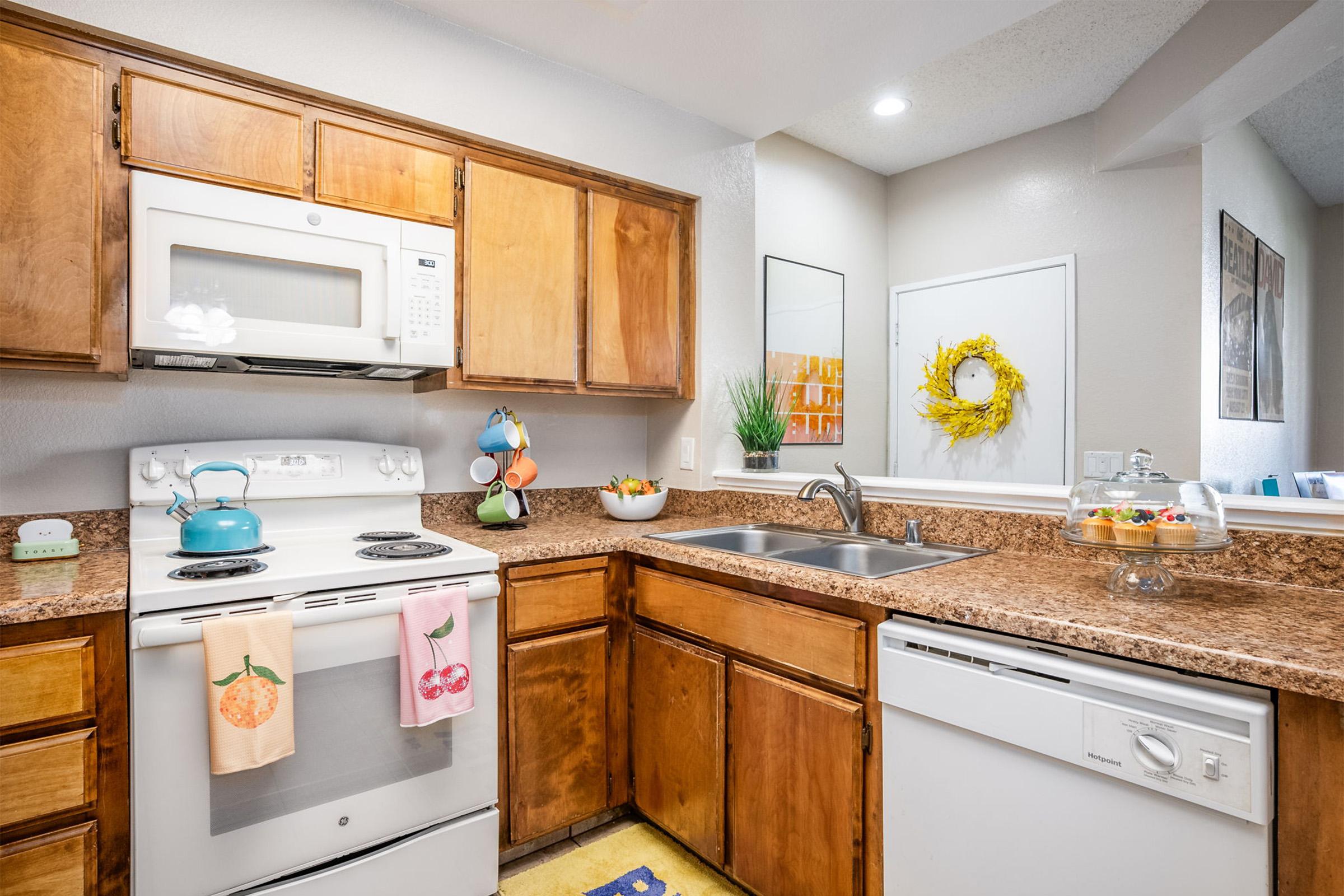 A cozy kitchen featuring wooden cabinets, a white stove and microwave, and a sink. The countertops are adorned with a fruit bowl and decorative items. A colorful dish towel hangs from the stove, and a small decorative wreath is mounted on the wall. The area is bright and inviting, reflecting a warm, homey atmosphere.
