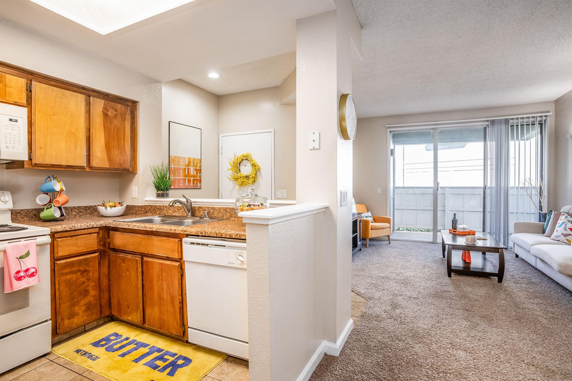 A cozy, well-lit apartment interior featuring a kitchen with wooden cabinets and granite countertops on the left, and a living area with a sofa, coffee table, and large window on the right. The kitchen includes a dishwasher, microwave, and decorative items, while the living area has colorful cushions on the sofa and a bright atmosphere.