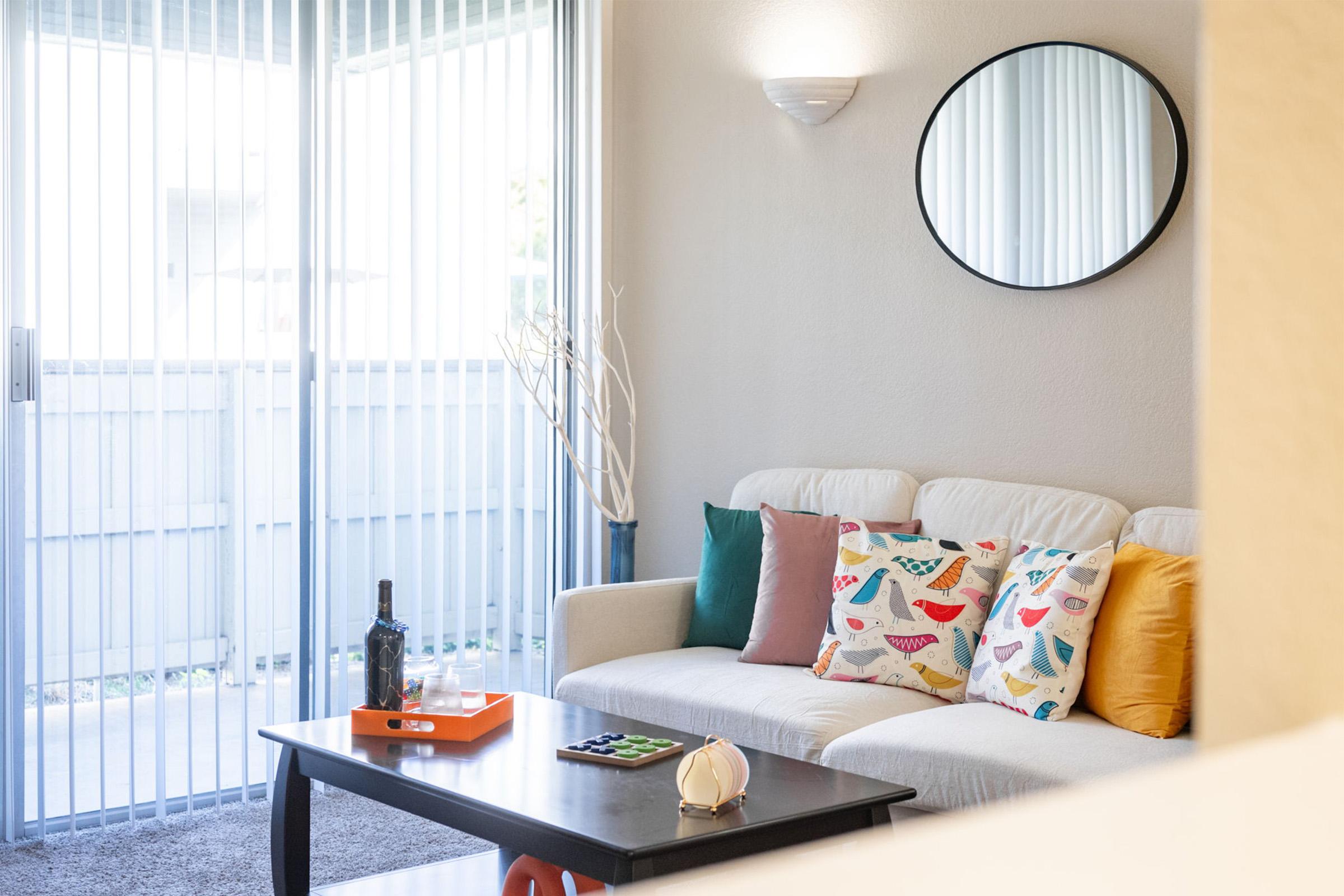 A cozy living room featuring a white sofa adorned with colorful patterned throw pillows. A black round mirror hangs on the wall, and there’s a wooden coffee table with a wine bottle, an orange tray, and a decorative item. Large sliding glass doors allow natural light to fill the space.