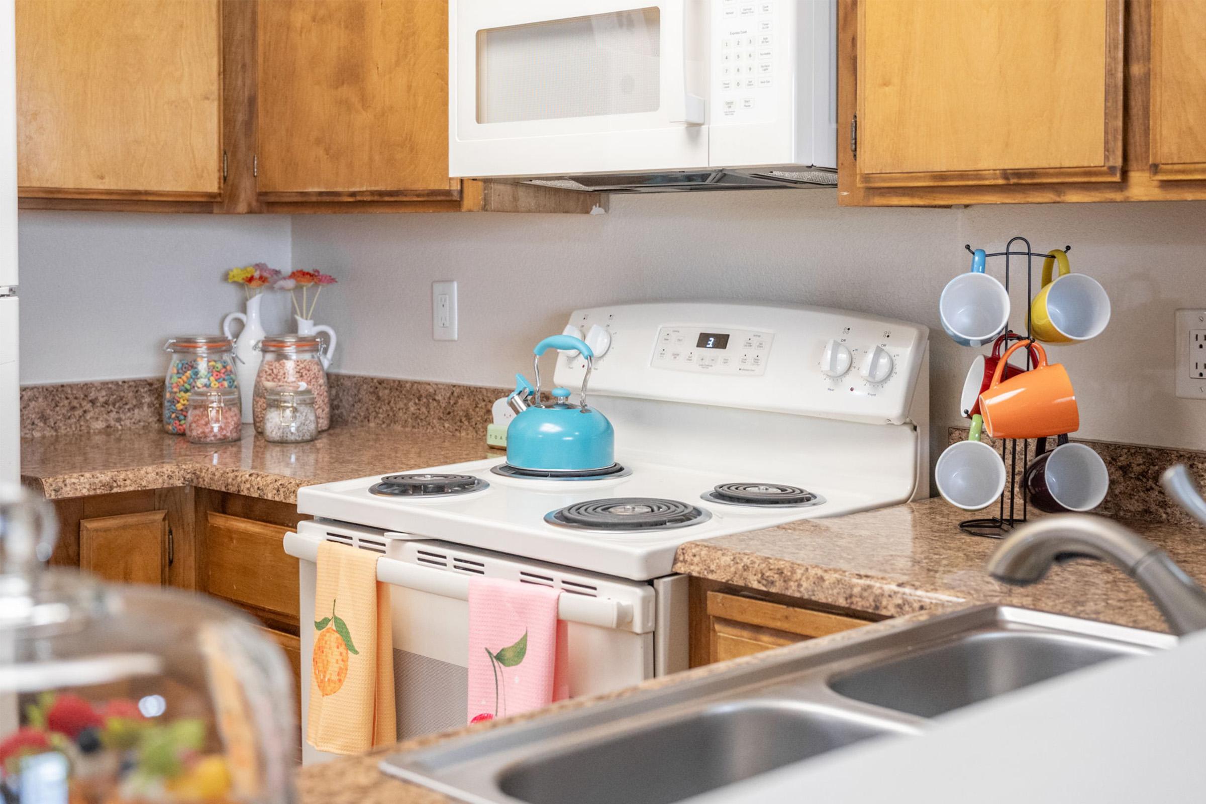 A cozy kitchen featuring wooden cabinets, a white stove with a blue kettle, and a microwave above. Colorful mugs hang on a rack, and there's a glass container displaying various candies. Two dish towels with fruit designs hang from the stove. A double sink is seen in the foreground.