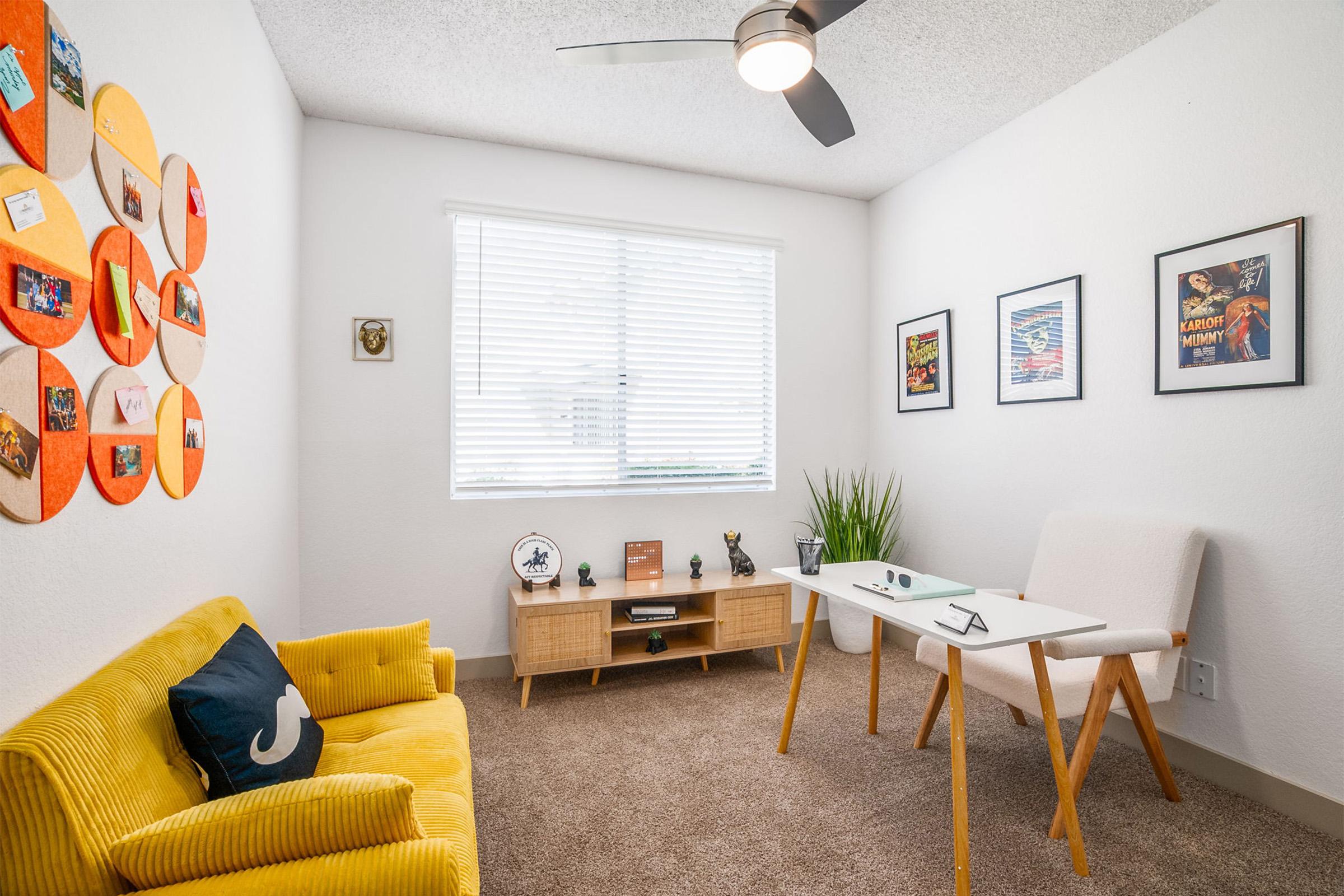A modern, well-lit office space featuring a yellow sofa, a wooden desk with a green notebook, and decorative artwork on the walls. There are framed pictures, a small plant, and an orange wall display with photographs. The room has a light-colored carpet and a ceiling fan.