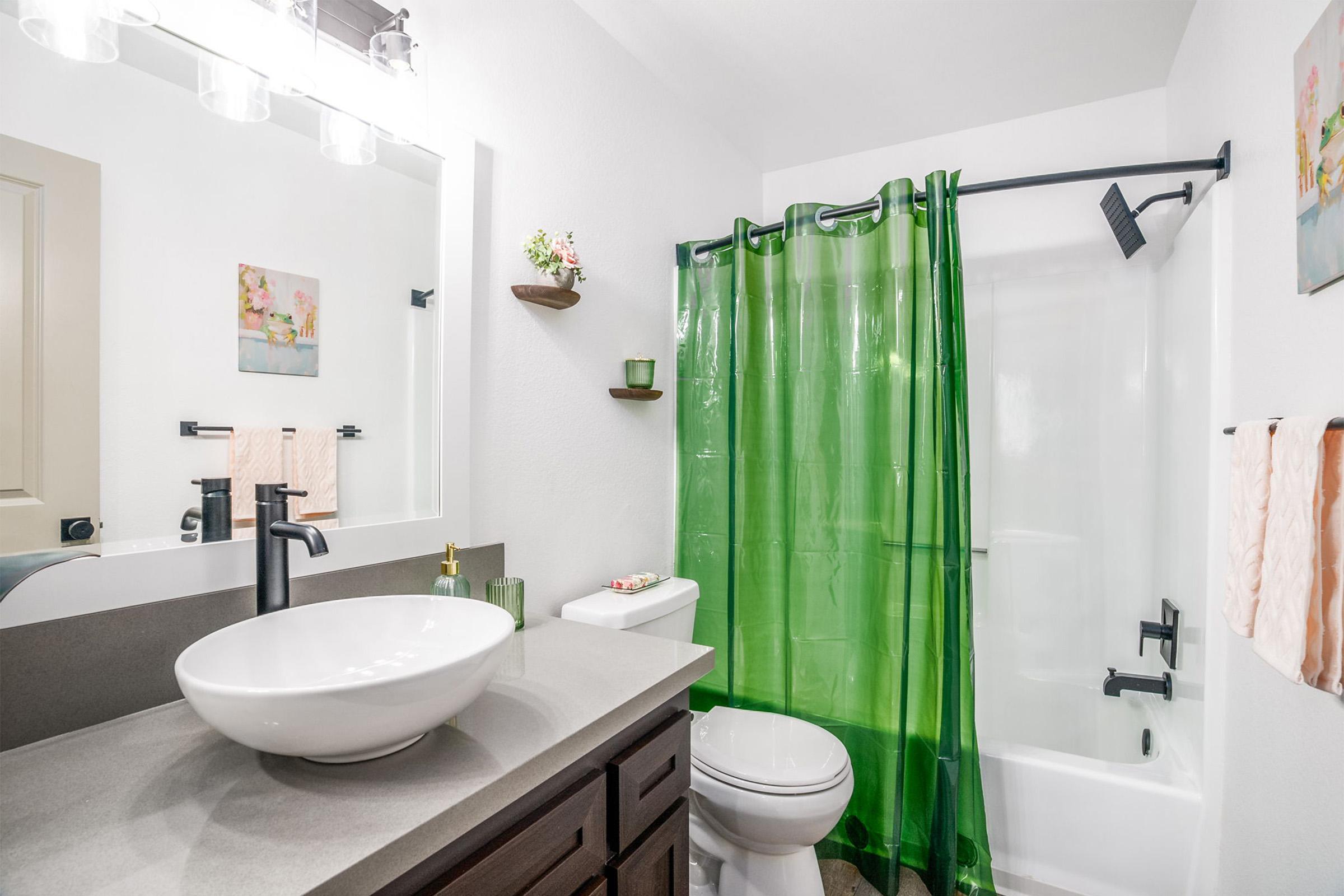 A bright and modern bathroom featuring a white countertop with a round sink, a toilet, and a bathtub with a green shower curtain. The walls are painted white, accented by small shelves with decorative items. Natural light is provided by a well-lit mirror above the sink.