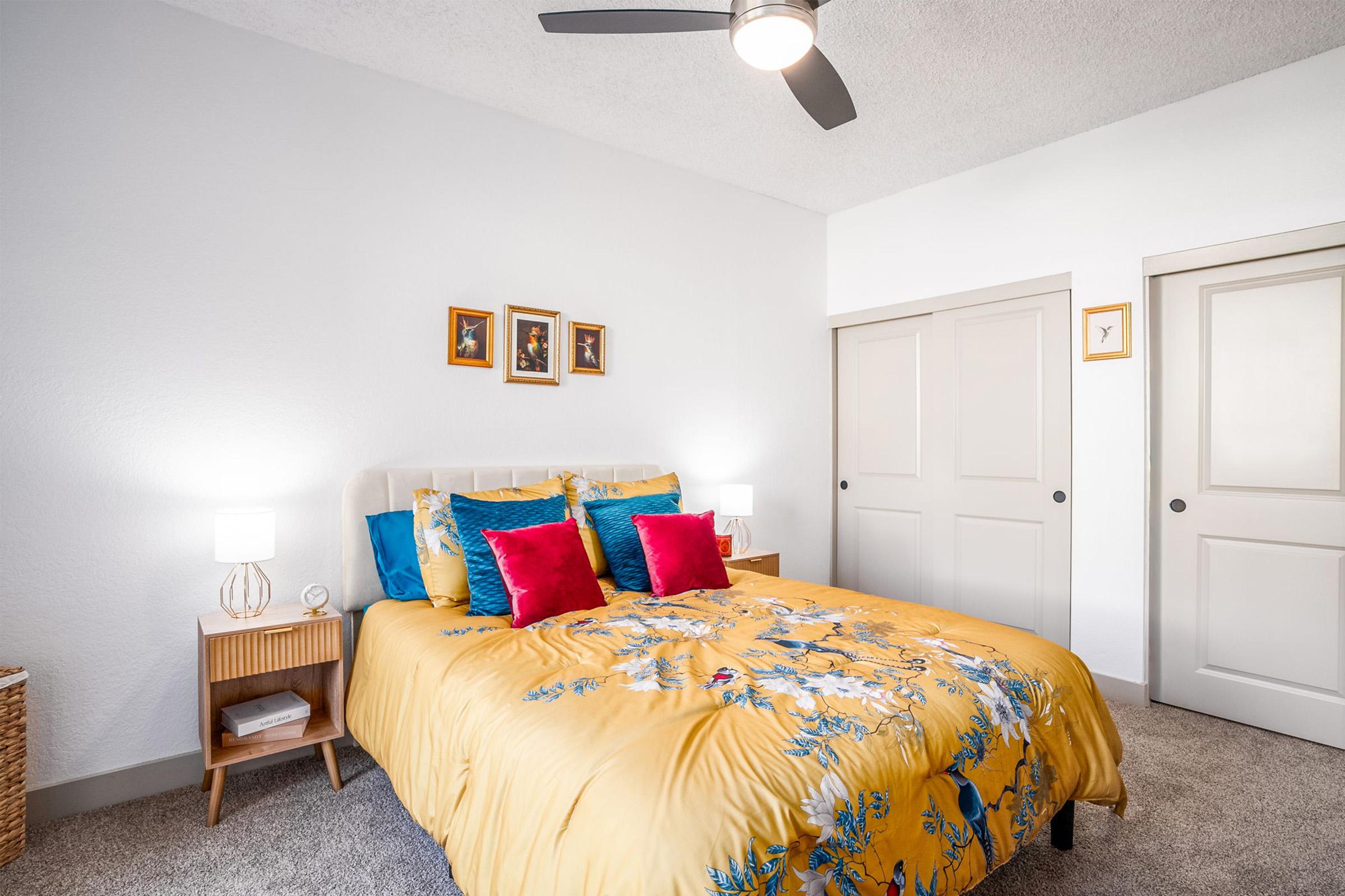 A cozy bedroom featuring a yellow floral-patterned bedspread with blue and red accent pillows. Soft lighting is provided by two bedside lamps on wooden nightstands. The walls are painted white, and there are framed pictures above the bed. A closet with sliding doors is visible in the background.
