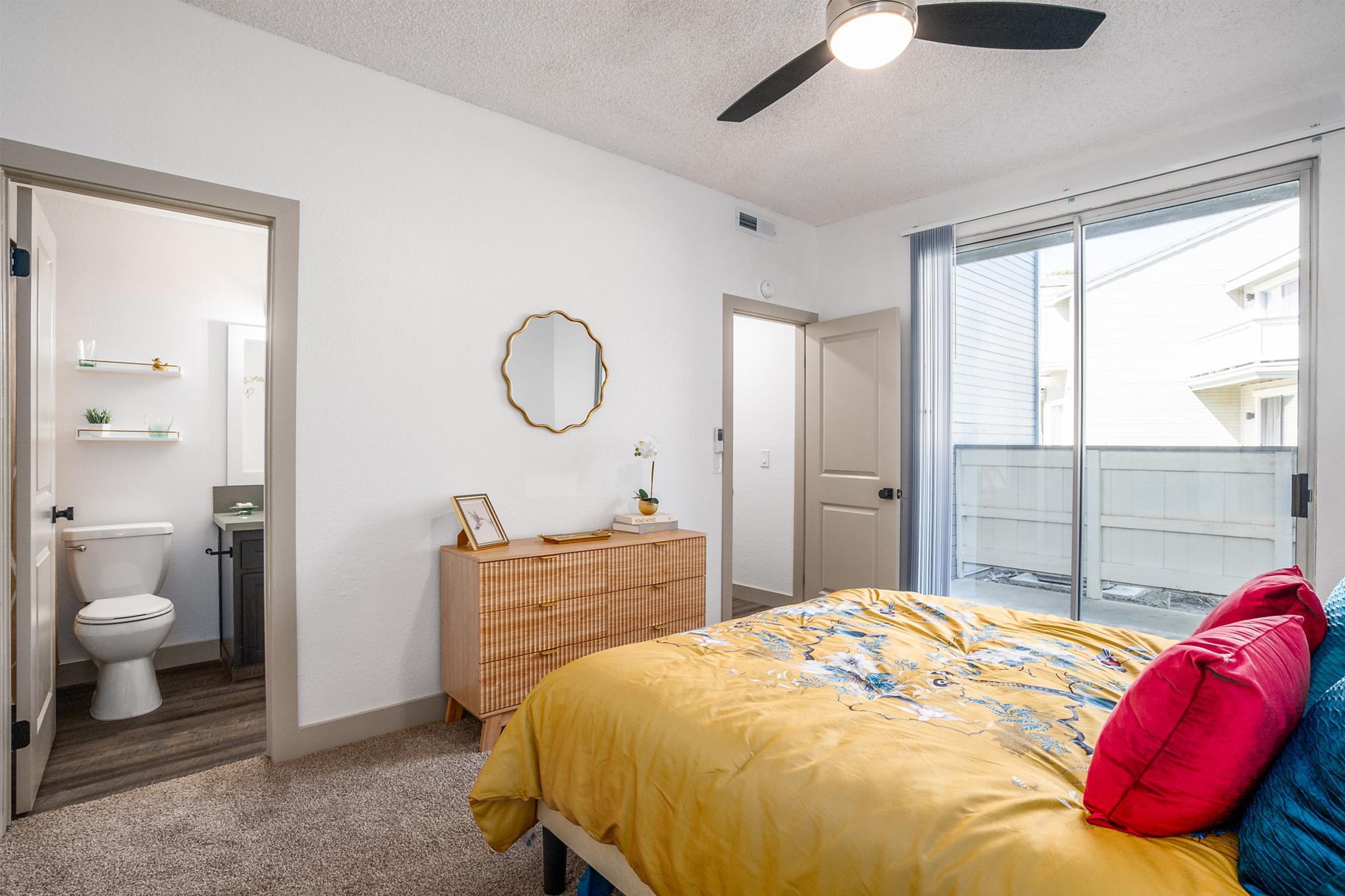 A cozy bedroom featuring a yellow bedspread with decorative pillows, a modern dresser, and a large window with sheer curtains. A door leads to a bathroom, partially visible, with a simple design. The room has neutral walls and carpet, creating a warm and inviting atmosphere.