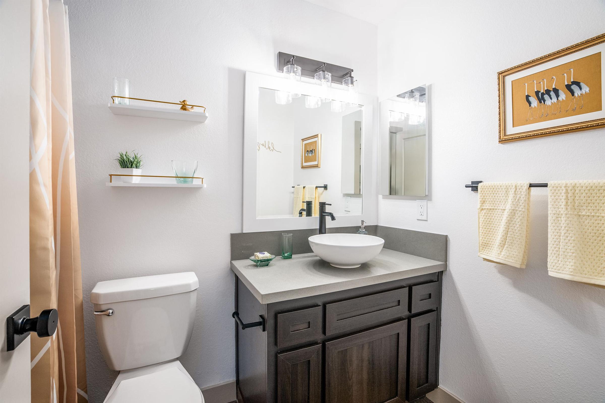 A modern bathroom featuring a toilet, a contemporary sink with a round basin, and a dark wood vanity. The space is well-lit with a mirror above the sink and stylish light fixtures. Shelves display decorative items and a framed artwork hangs on the wall. Soft yellow towels are neatly arranged.