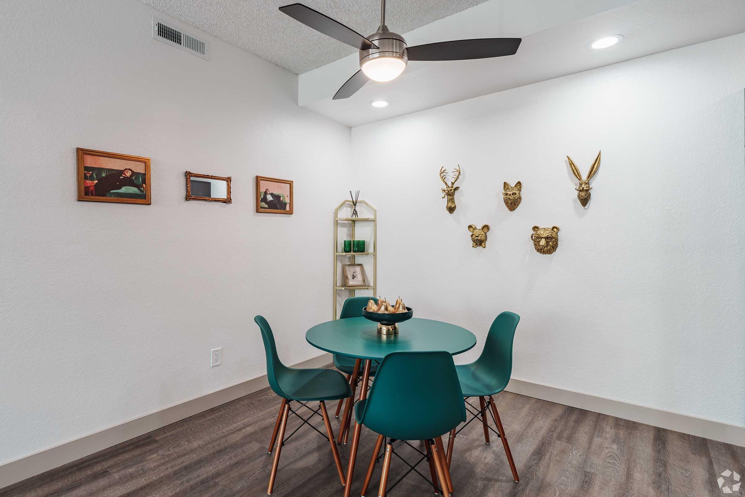 A modern dining area featuring a round green table surrounded by four teal chairs. On the walls, there are framed art pieces and mounted animal heads, including a deer and other decorative masks. A stylish shelf holds small green plants and decor items. The room has a ceiling fan and white walls.