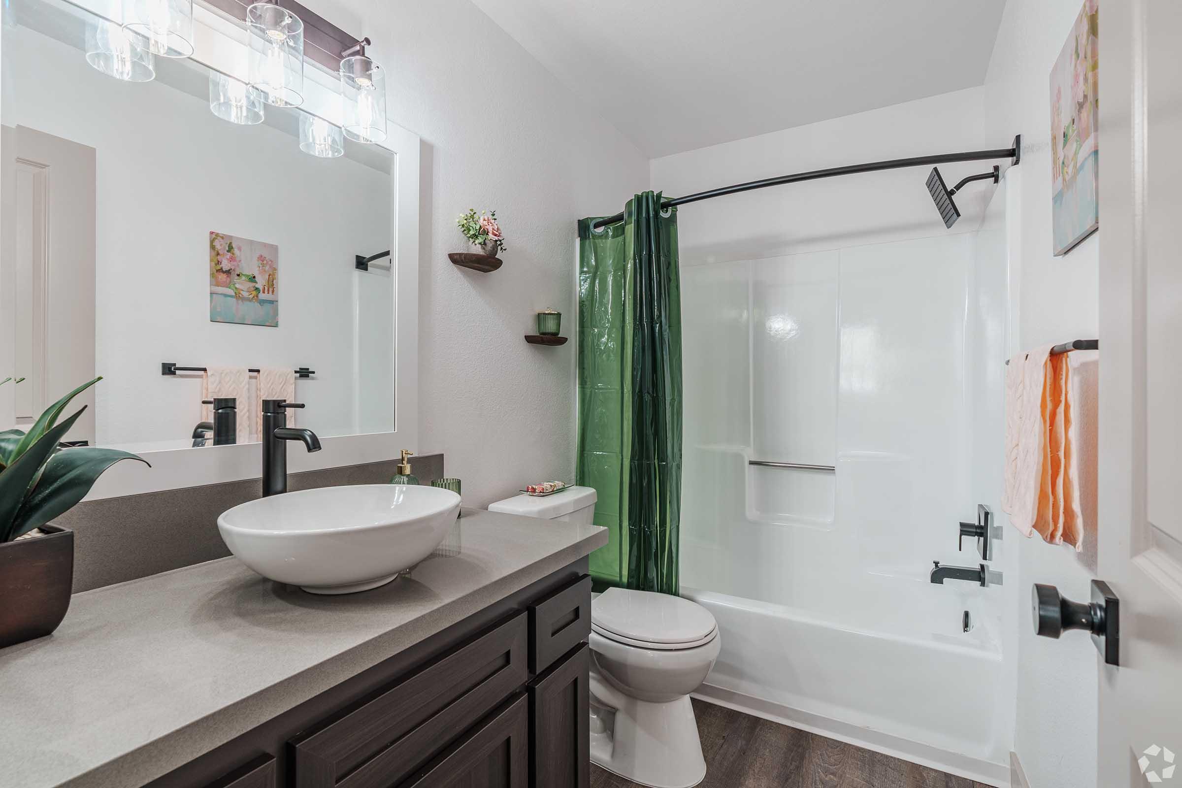 A modern bathroom featuring a white bathtub with a glass shower curtain, a wall-mounted sink with a round basin, and dark cabinetry. The space includes greenery accents, decorative wall art, and two towels hanging on a rack. A large mirror with stylish lighting completes the contemporary design.