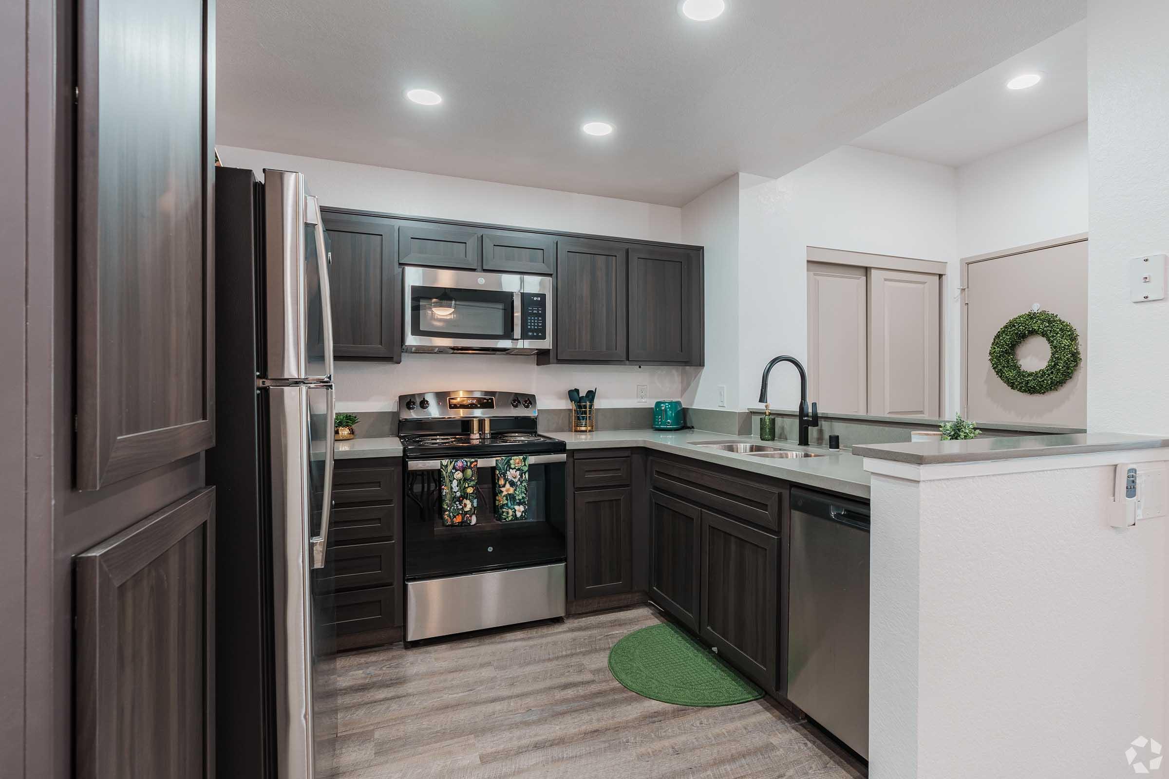 Modern kitchen with dark cabinets, stainless steel appliances, and a spacious countertop. Features include a microwave, stove, and sink with a faucet. Green decor accentuates the space, including a small rug and a wreath on the wall. Good lighting from recessed ceiling fixtures enhances the overall ambiance.