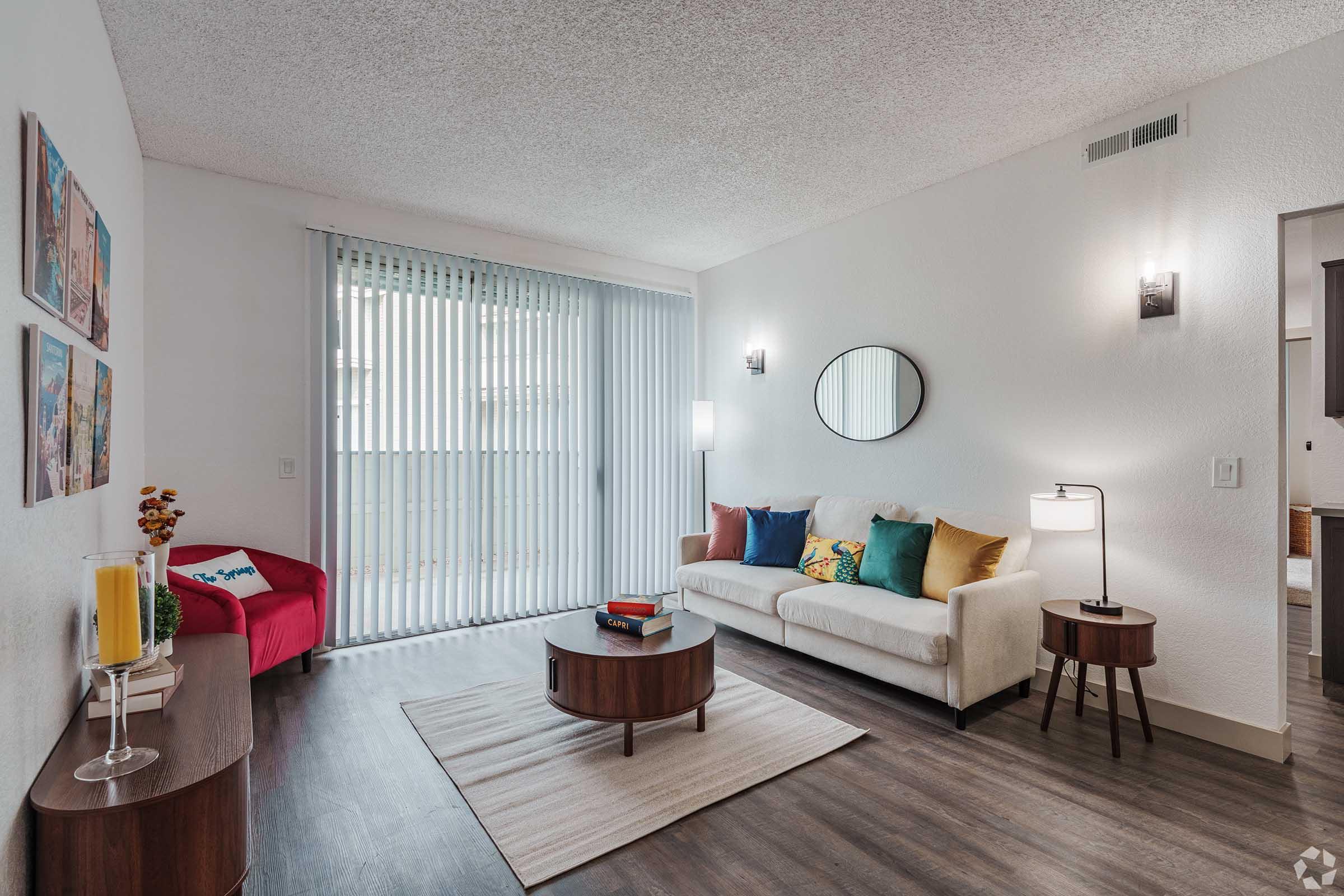 A bright living room featuring a light-colored sofa with colorful pillows, a round wooden coffee table, and a modern floor lamp. There is a red chair, a decorative vase with flowers, and a large mirror on the wall. Vertical blinds cover a sliding glass door, letting in natural light.