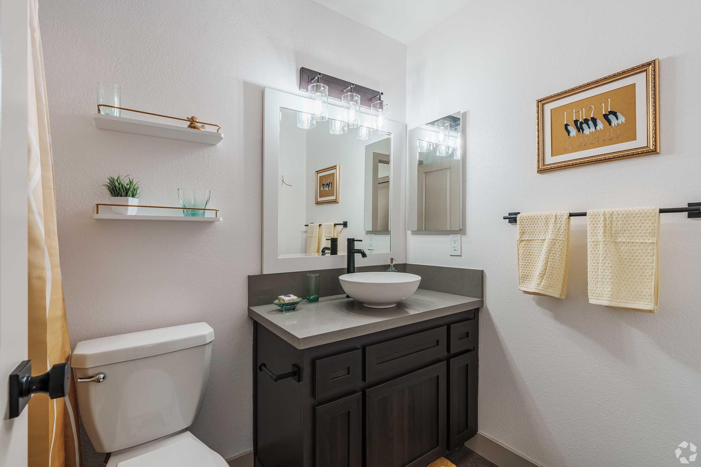 A modern bathroom featuring a sleek design with a round vessel sink on a dark vanity, a toilet, and wall-mounted shelves. The walls are painted a light color, and there are glass light fixtures above the mirror. A towel rack with a yellow towel adds a pop of color to the space.