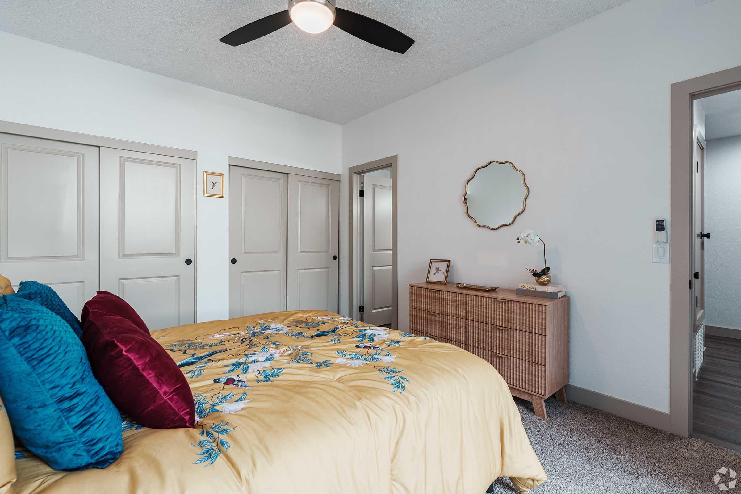 Cozy bedroom featuring a yellow embroidered comforter, vibrant blue and burgundy pillows. There’s a wooden dresser with decor and a wall mirror. Two closed closet doors and a door leading to another room are visible, along with a ceiling fan for added comfort. The decor is modern and inviting.