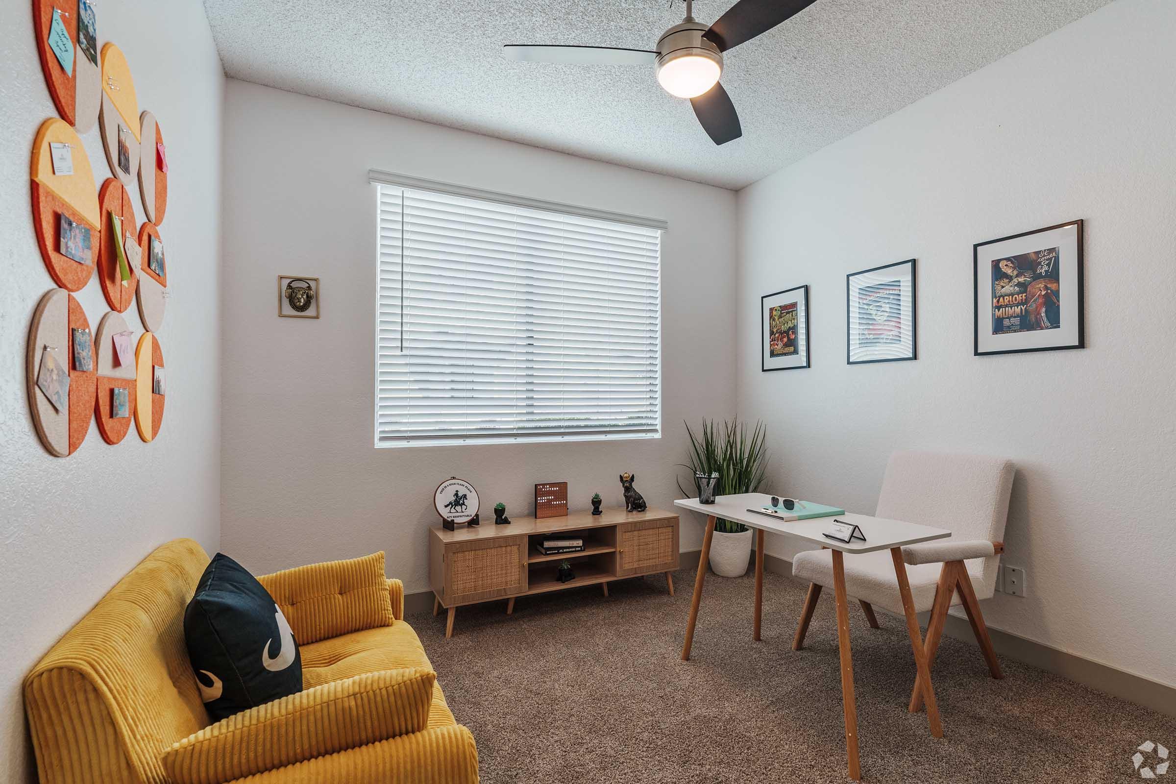 A cozy, well-lit small room featuring a yellow couch, a stylish desk with a chair, and a wall adorned with colorful circular art pieces. The room includes a window with white blinds, framed pictures on the wall, and a green plant, creating a bright and inviting atmosphere.