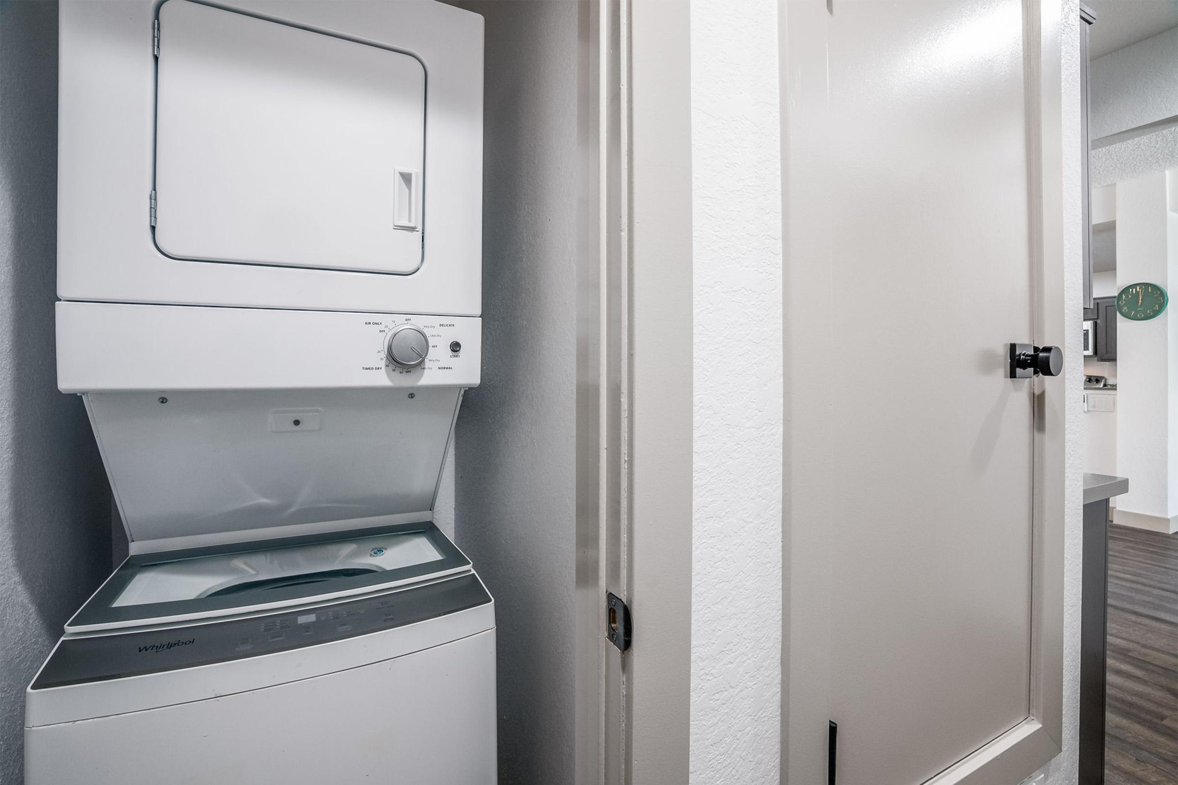 A stackable washer and dryer unit in a small laundry space next to a closed door. The walls are painted a light color, and the flooring is a dark, wood-like material. A clock can be faintly seen in the background, indicating a kitchen area.