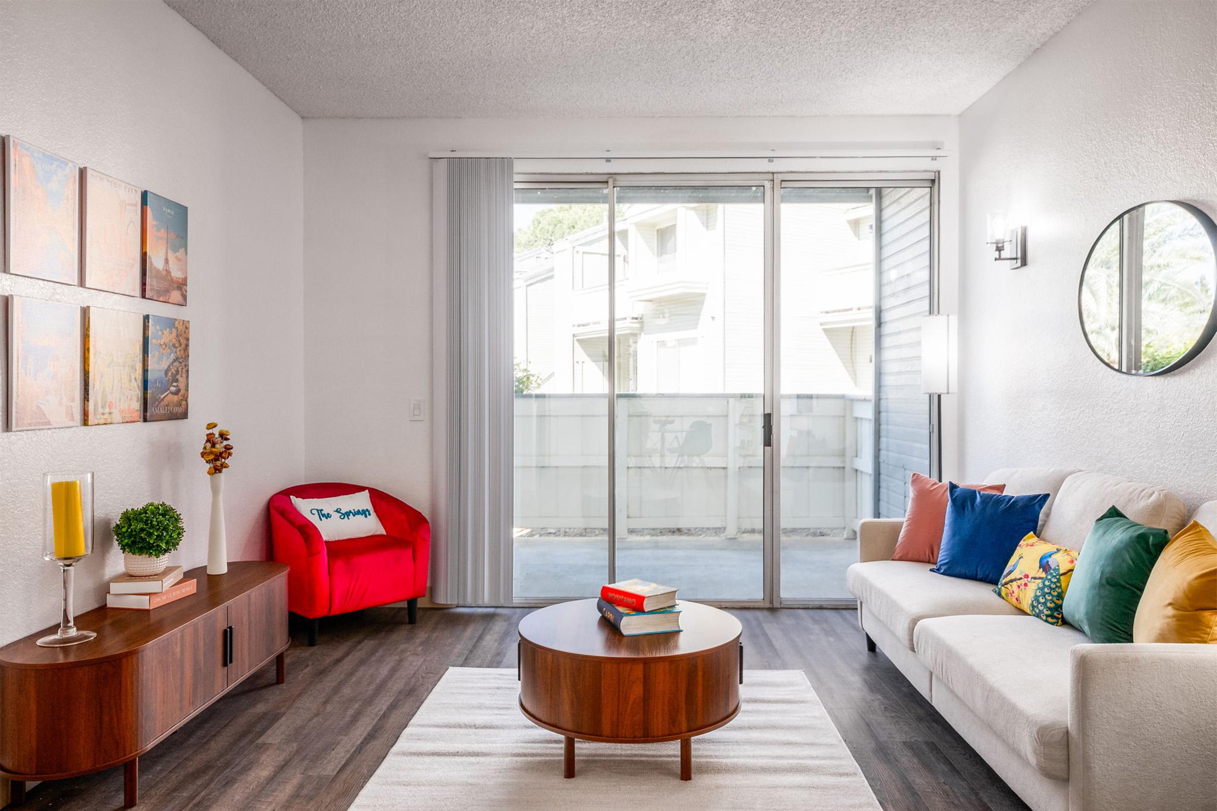 A cozy living room featuring a white sofa adorned with colorful cushions, a round coffee table, and a stylish side table with a lamp. There's a red accent chair, decorative wall art, and large sliding doors leading to a balcony. Soft natural light fills the room, creating a welcoming atmosphere.