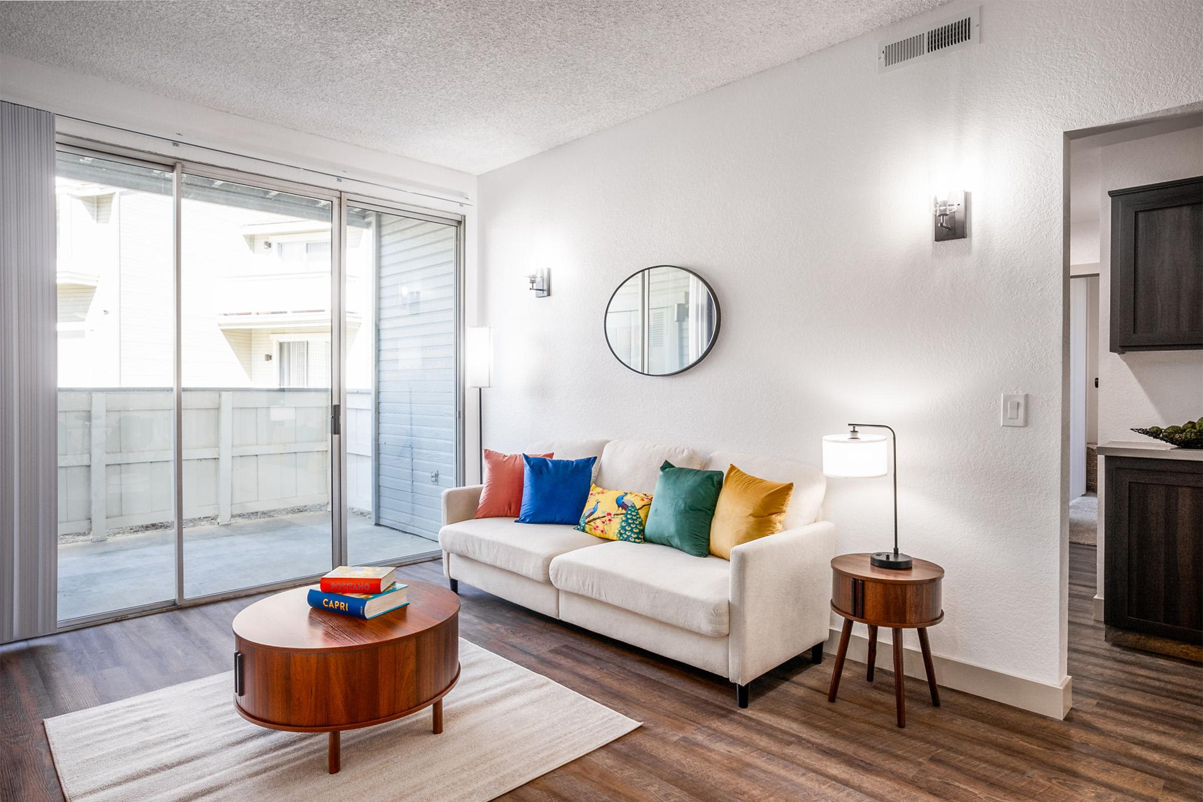 Bright and modern living room featuring a white sofa adorned with colorful pillows, a round wooden coffee table with books, and a stylish floor lamp. Large sliding glass doors offer access to a balcony, while a circular mirror and elegant wall sconces add a touch of sophistication to the space.
