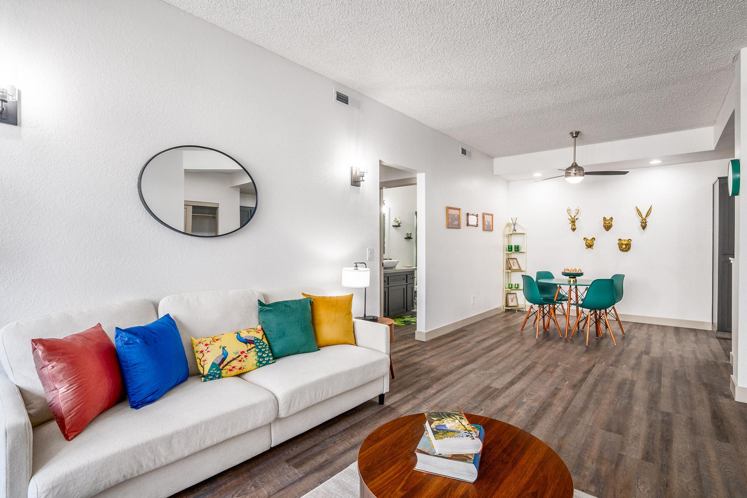 A modern living room featuring a white sofa with colorful cushions, a round mirror on the wall, a wooden coffee table, and light-colored flooring. In the background, there’s a small dining area with green chairs, a display of decorative items, and art on the walls, creating a stylish and inviting atmosphere.